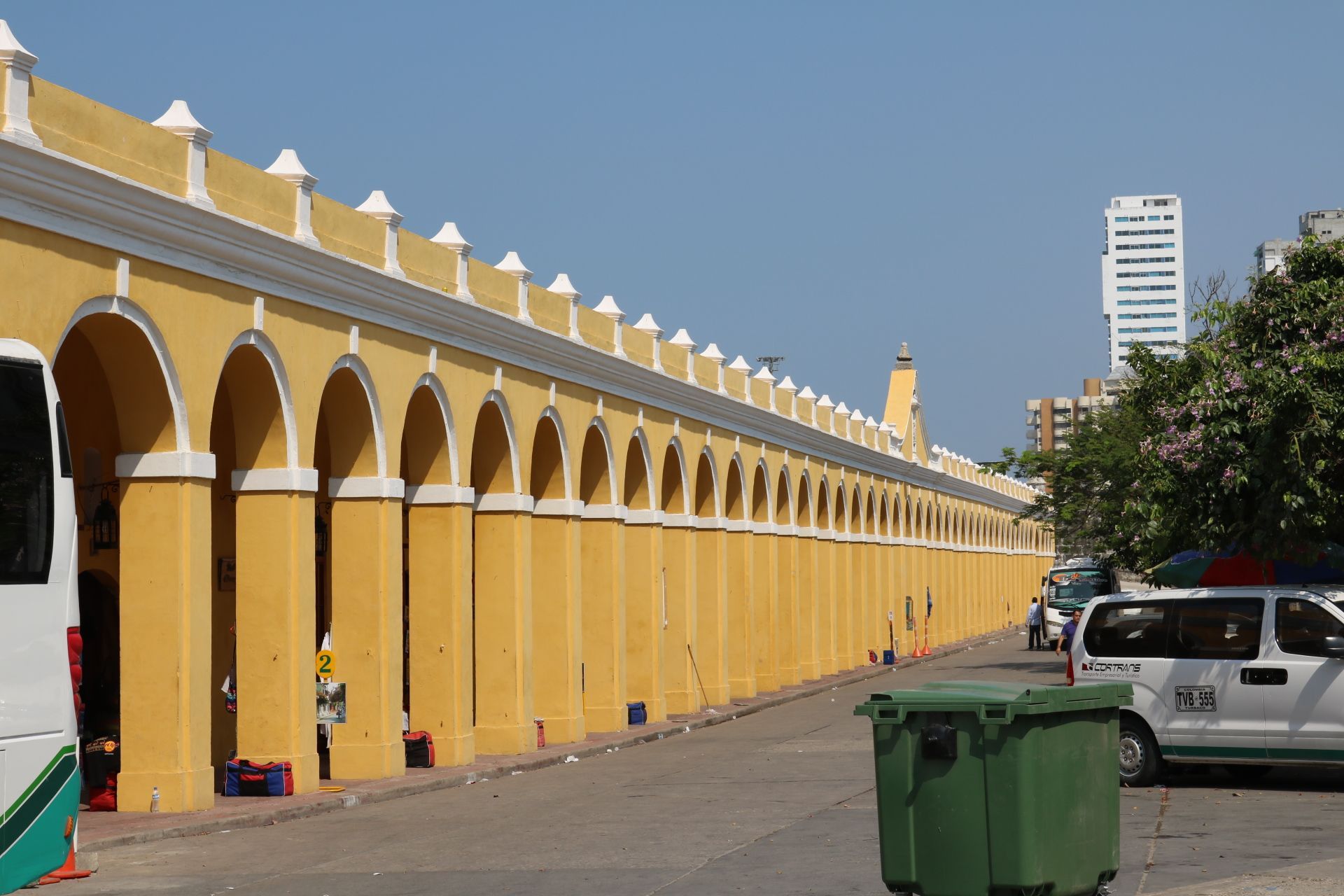 Yellow arched wall along a street with parked cars and green trash bins under a clear sky