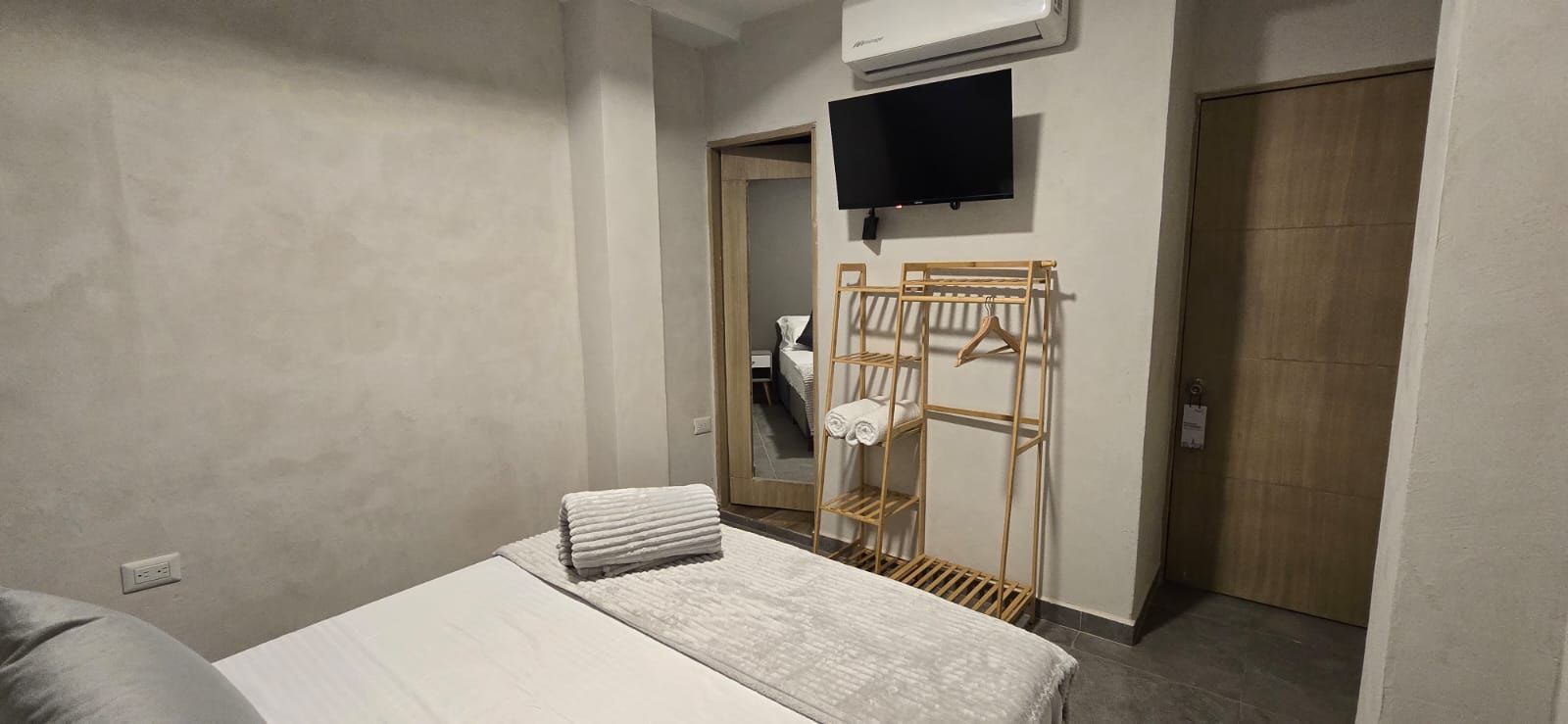 Small hotel room with bed against brick wall, desk, chair, and clothes rack.