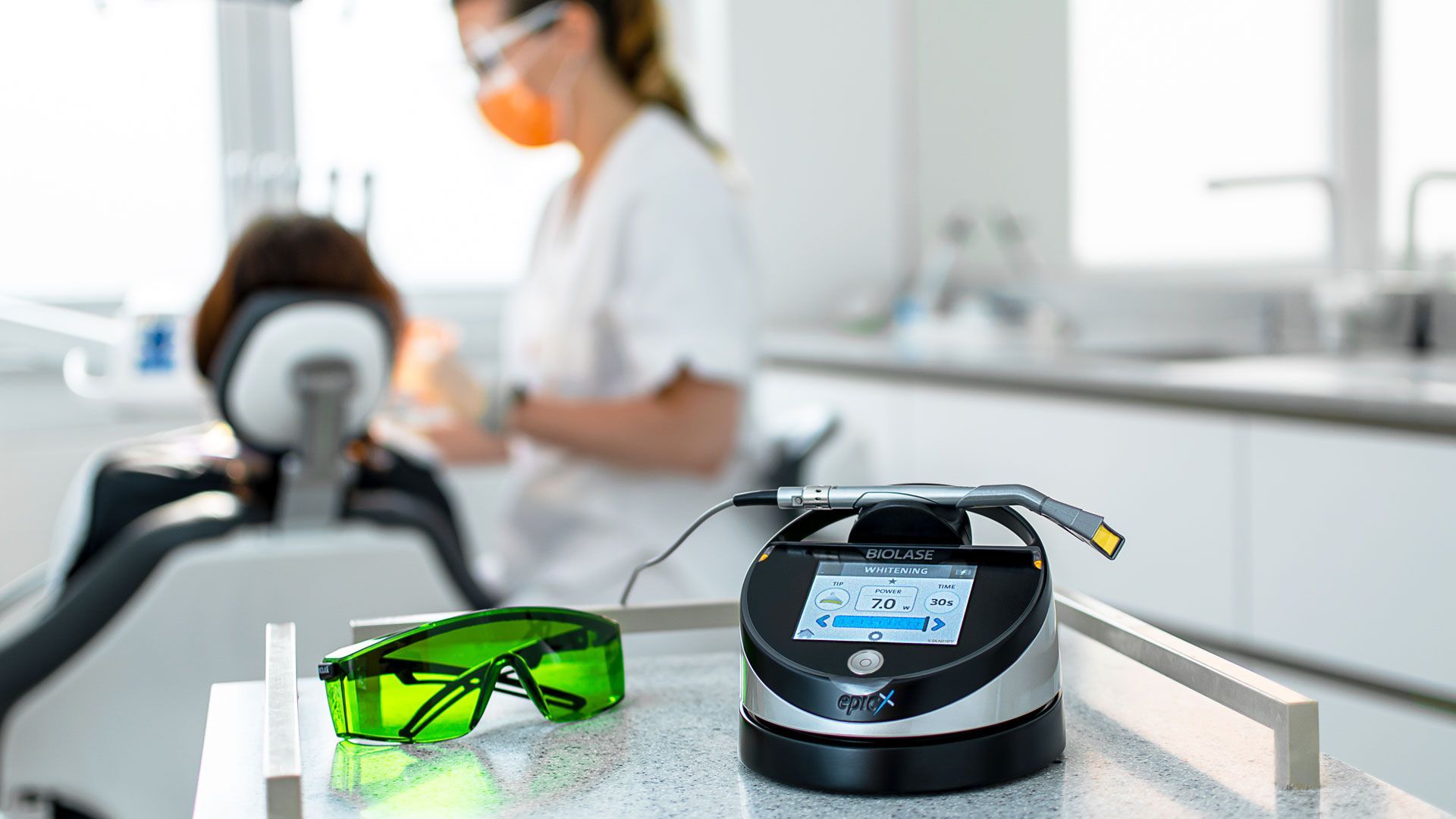 Biolase Epic X Gerät für Laser-Zahnaufhellung und Schutzbrille auf dem Tisch, im Hintergrund eine Patientin in Behandlung