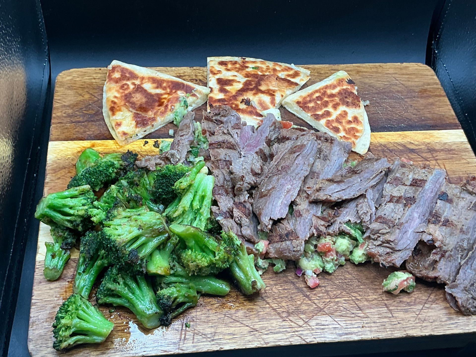 Grilled steak, broccoli, and flatbread on a wooden cutting board, dark background.