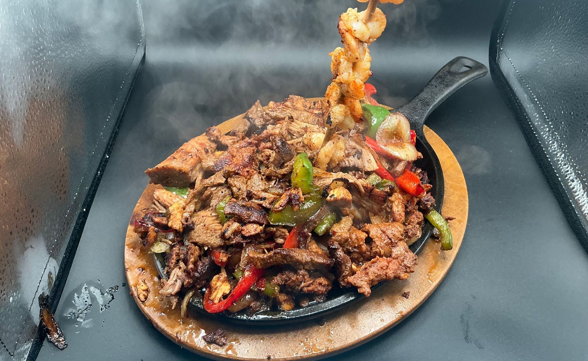 Sizzling fajita platter with meat and vegetables, topped with shrimp skewers; steam rising.