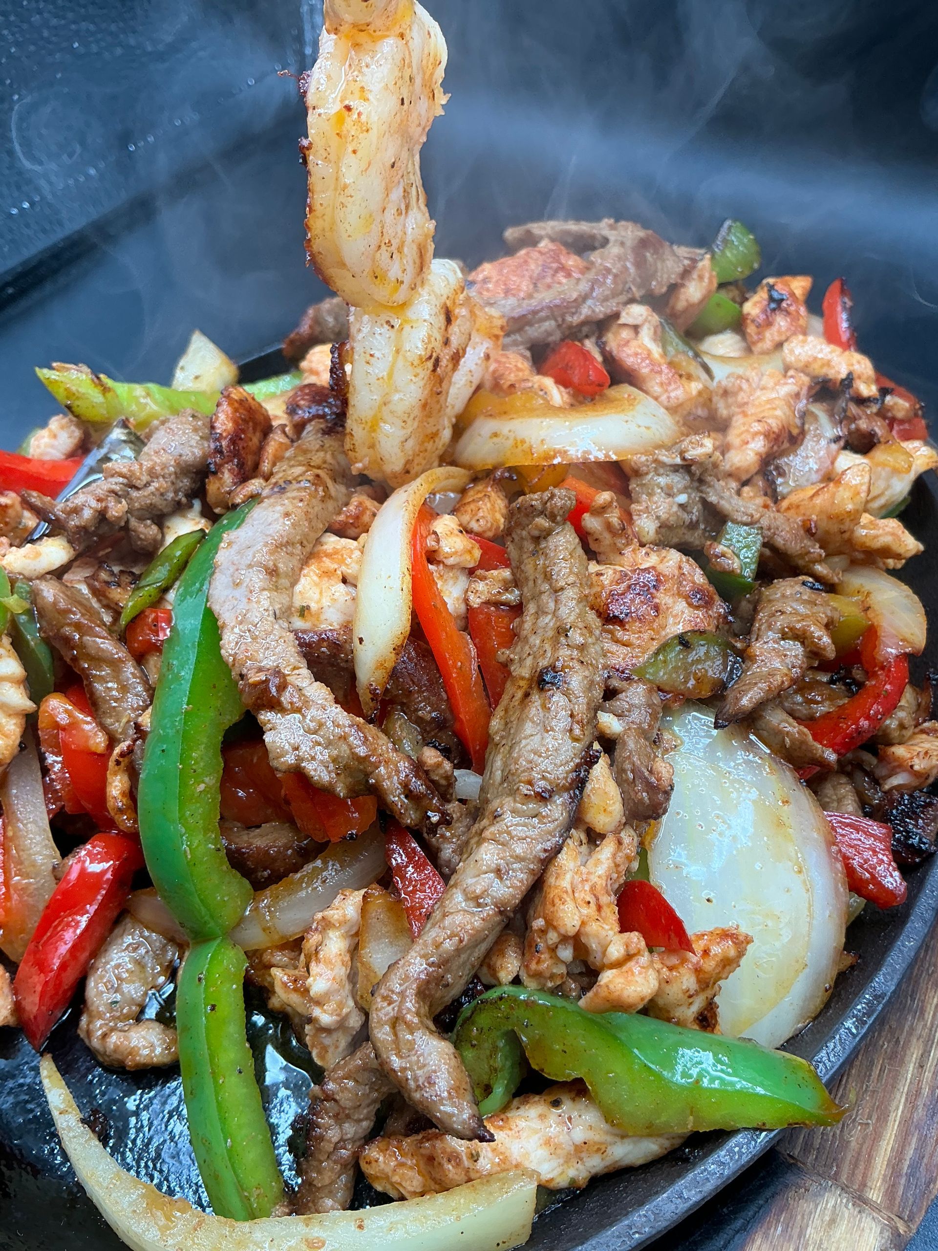Sizzling fajitas with steak, chicken, shrimp, peppers, and onions in a hot cast iron skillet.