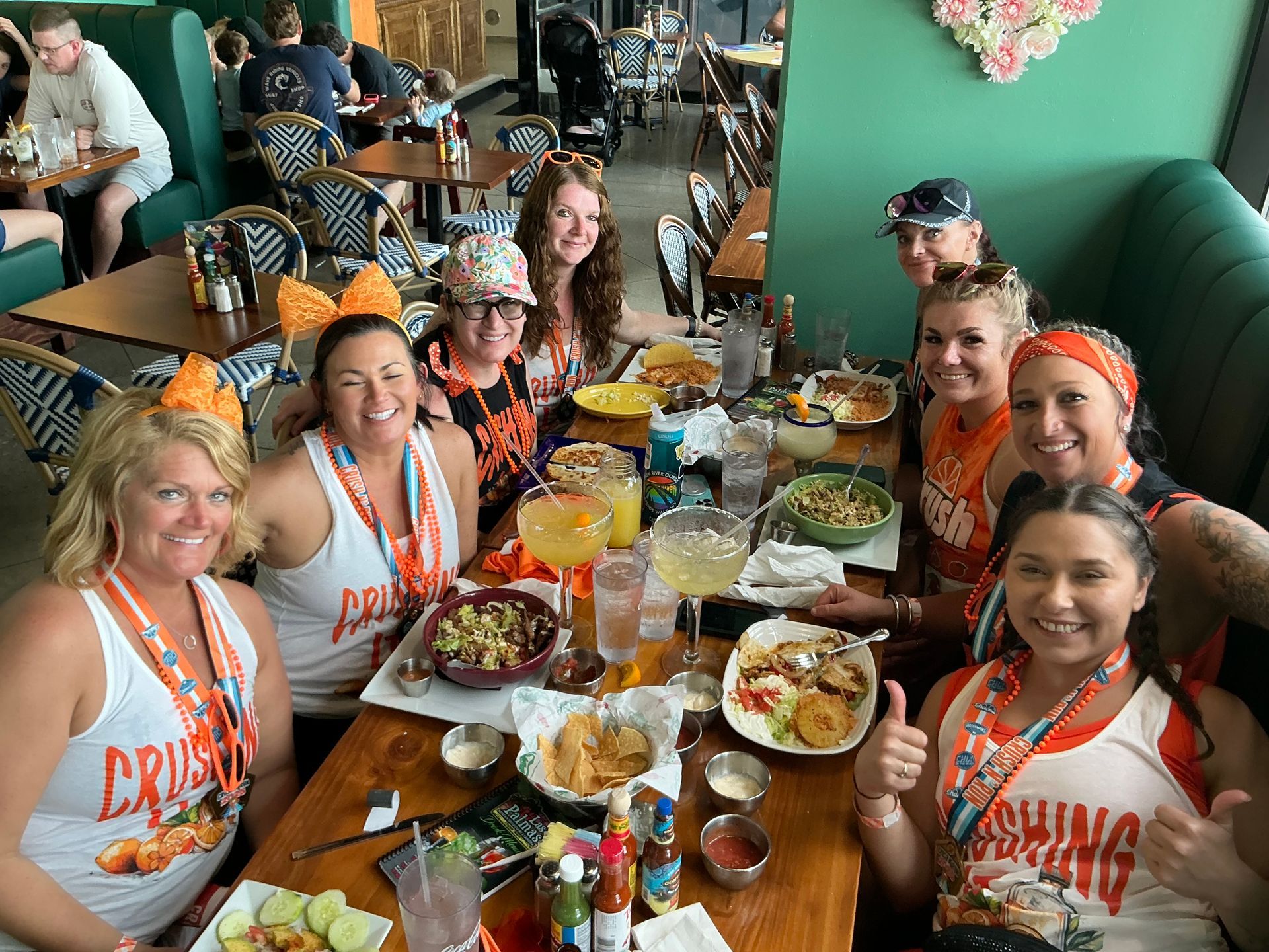 A group of nine people at a restaurant, smiling and eating food. They wear matching orange shirts and are at a table.