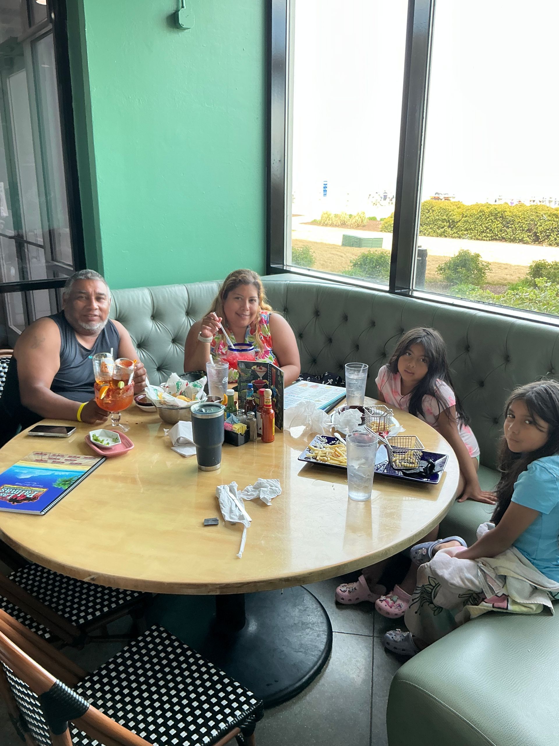 Family eating at a restaurant by a window, one is smiling, food on the table.