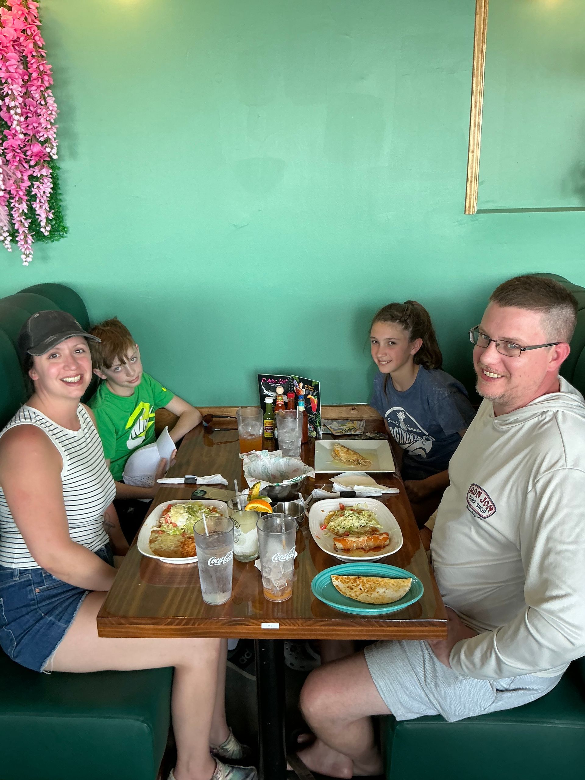 Family of four seated at a table in a restaurant, eating. Green wall, flowers, and food on plates are visible.
