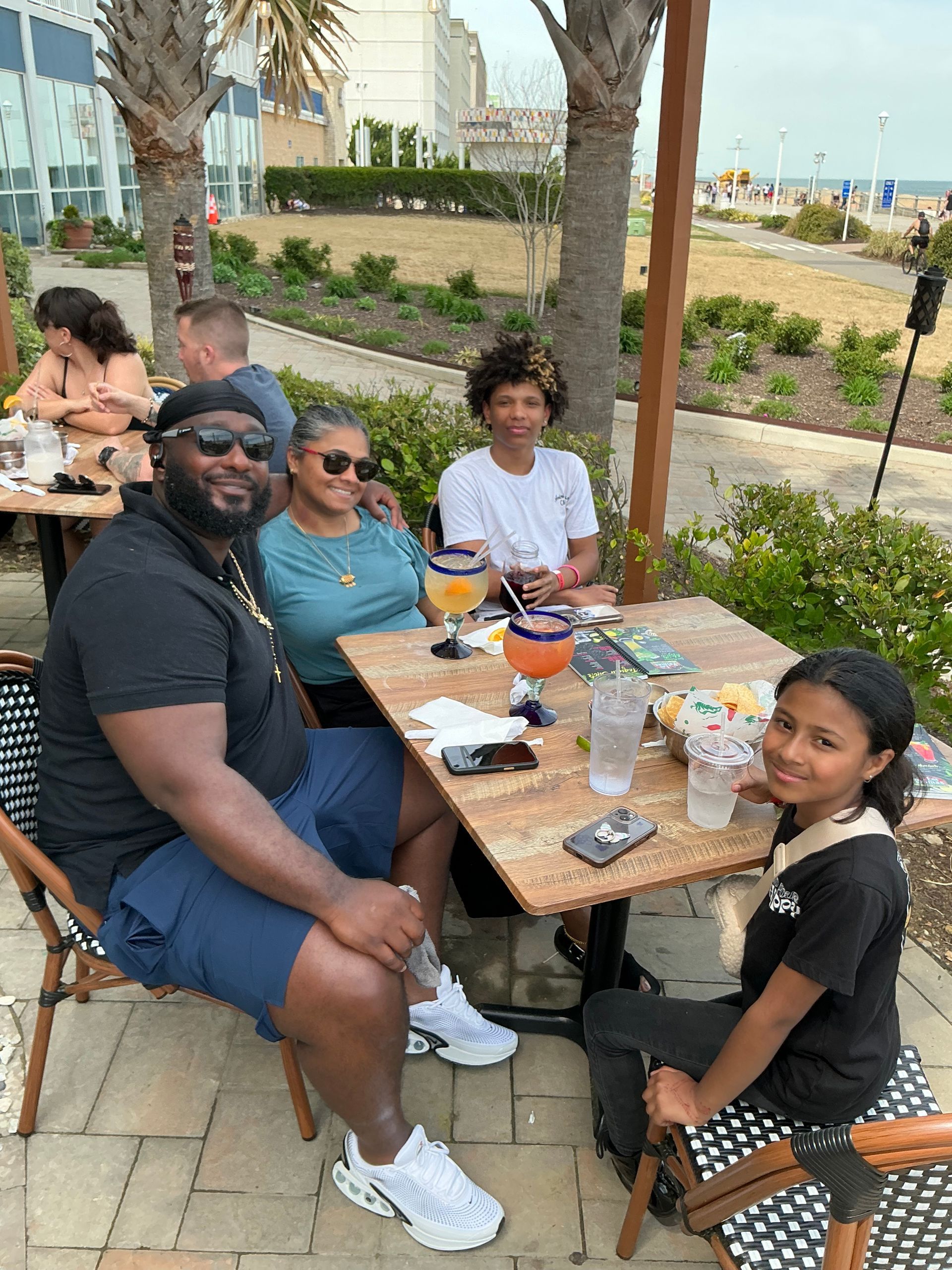 Family at outdoor cafe, ocean view. They are smiling, drinking, sitting at a wooden table, under a wooden structure.