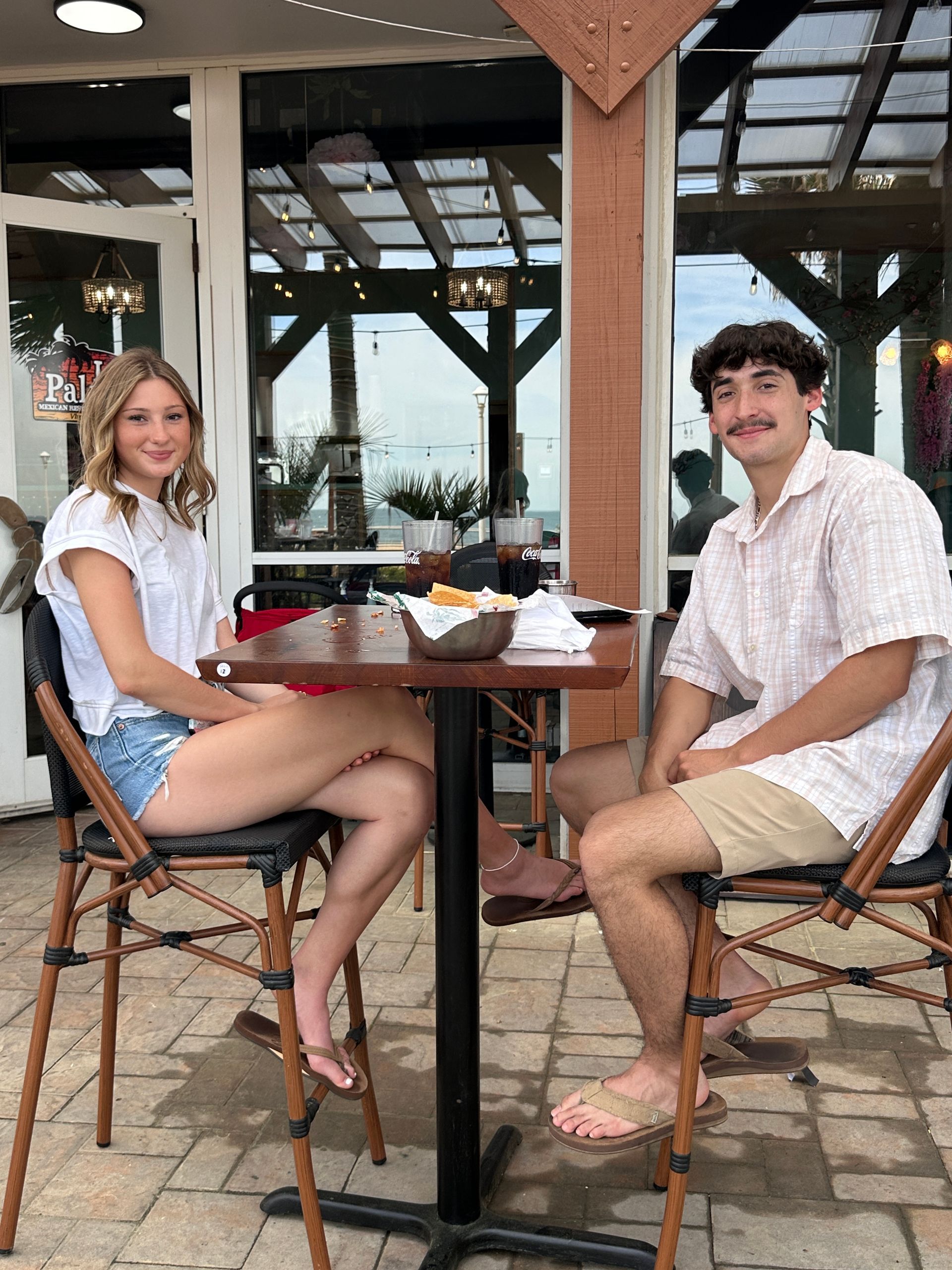 A couple sits at a table outdoors. The woman wears a white top and denim shorts; the man wears a patterned shirt and khaki shorts.