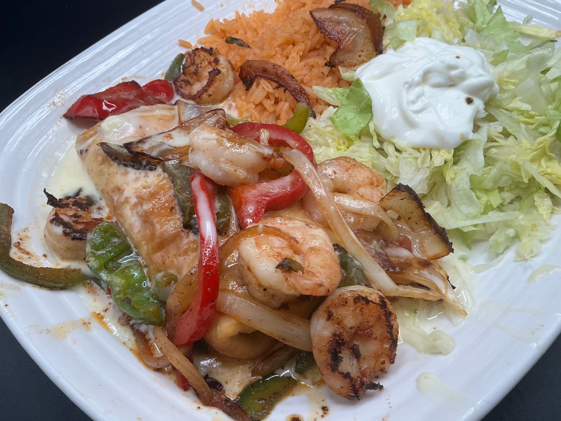 Seafood fajitas with shrimp, scallops, bell peppers, onions, rice, sour cream, and lettuce on a white plate.