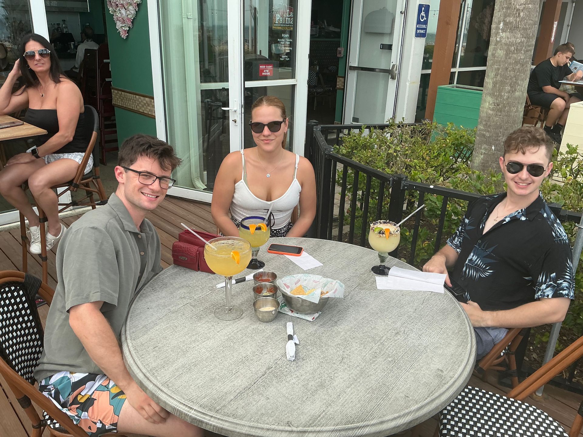 Three people at outdoor table with drinks, smiling. Woman in white top, two men. Exterior restaurant setting.