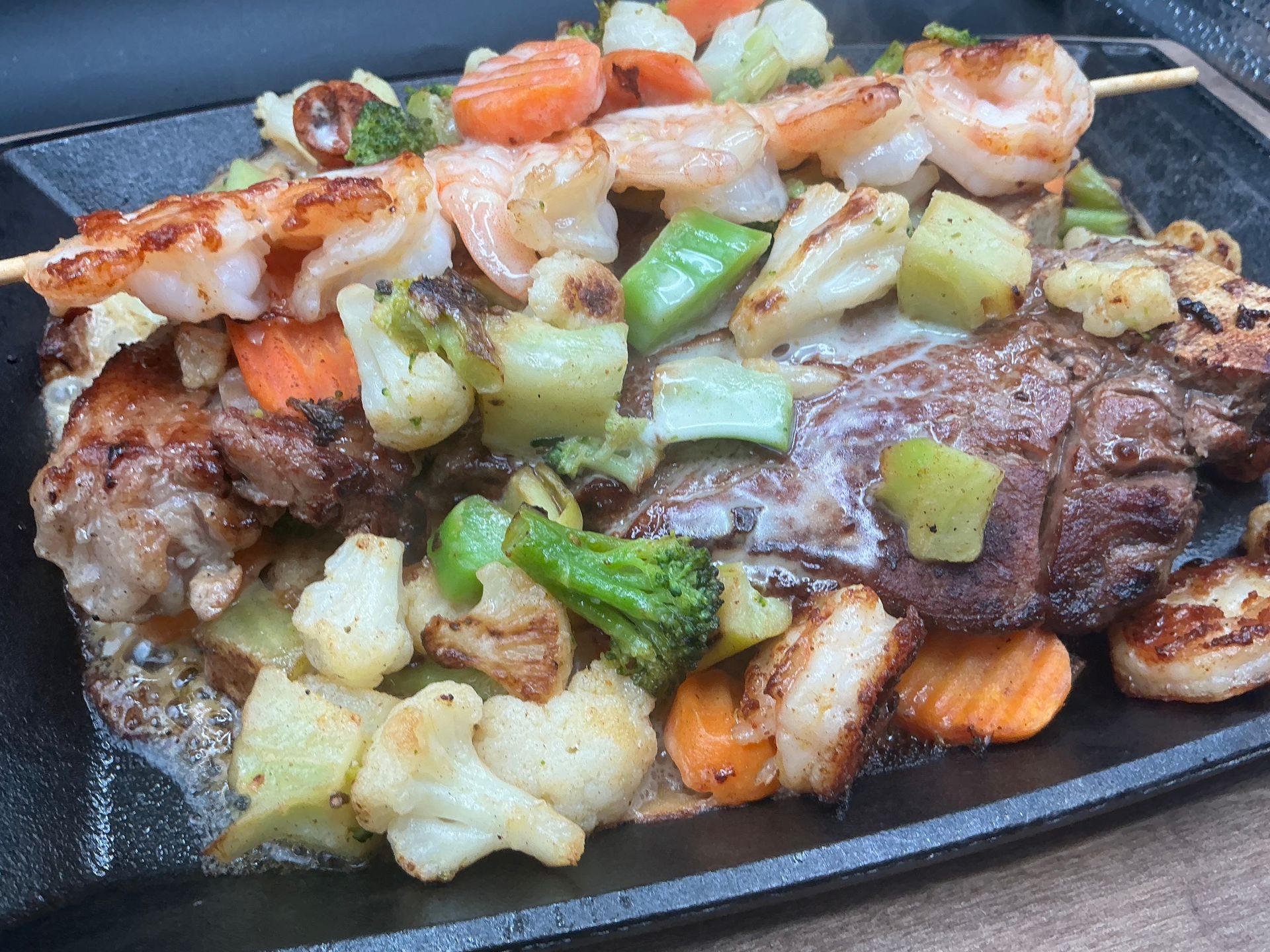Grilled meat and shrimp skewers with vegetables on a black platter.