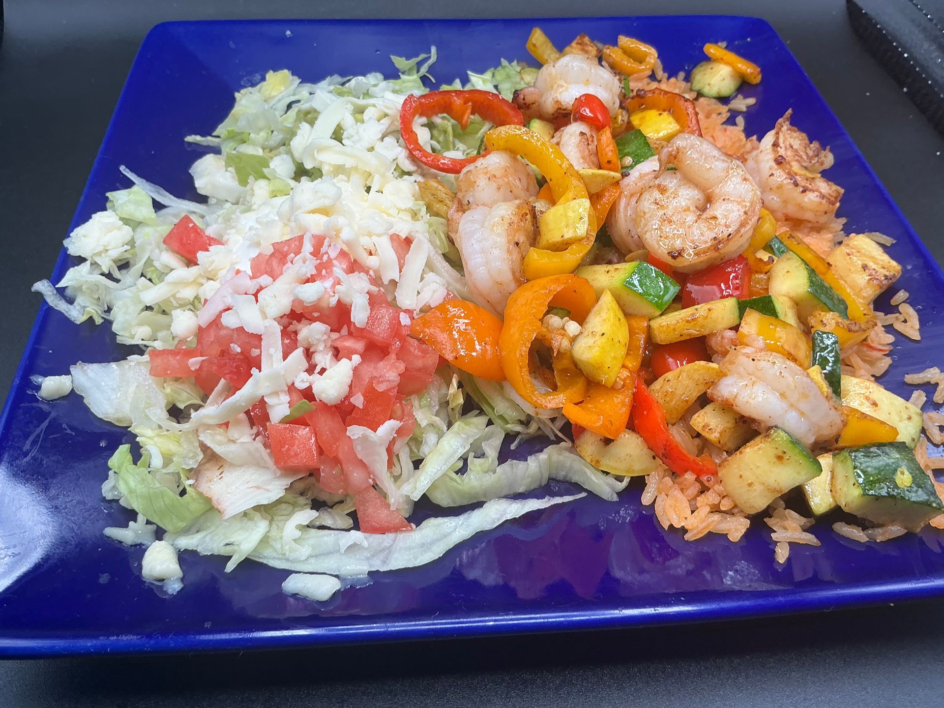 Shrimp fajitas on a blue plate: shrimp, peppers, zucchini, rice, lettuce, tomato, and cheese.