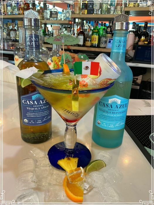 Margarita in a large glass with Casa Azul tequila bottles. Garnish with fruit, Mexican flag. Bar setting.