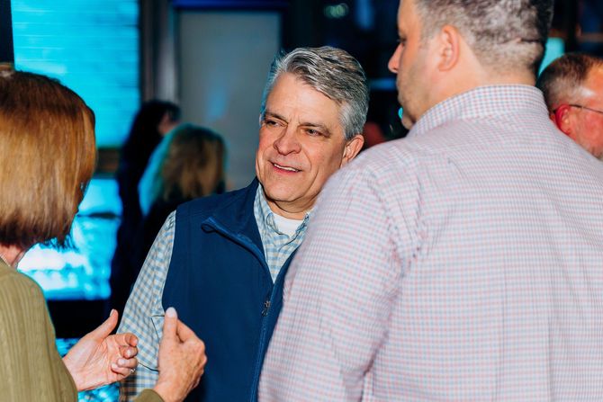Two men at an event, one eating from a plate, smiling. Others are blurred in the background.