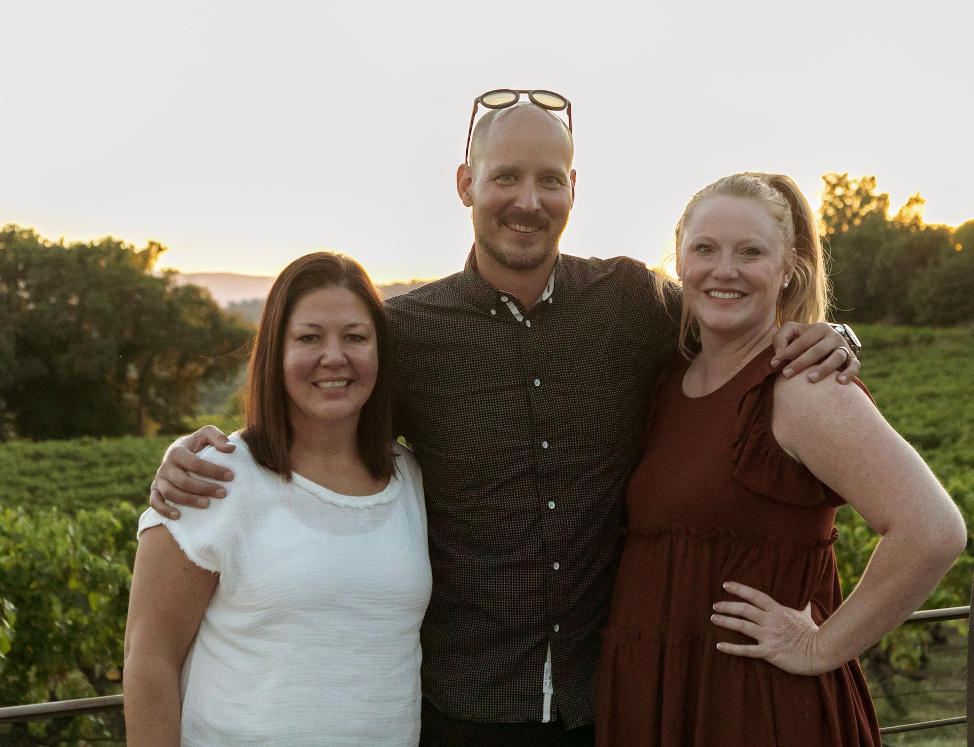 Three people smiling at sunset in a vineyard; man with arms around women's shoulders.
