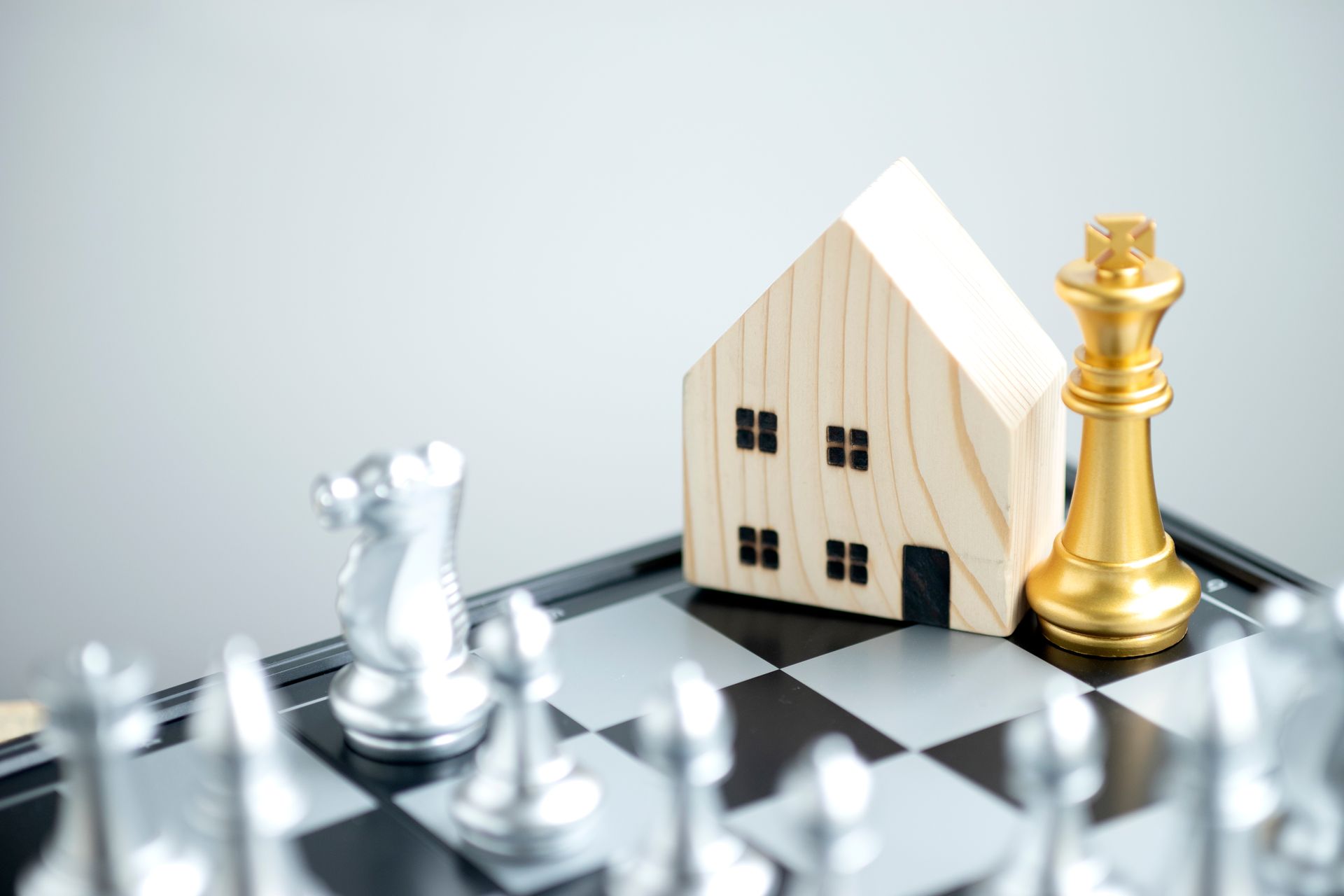 Chessboard with toy house and gold king piece. Silver chess pieces in foreground.