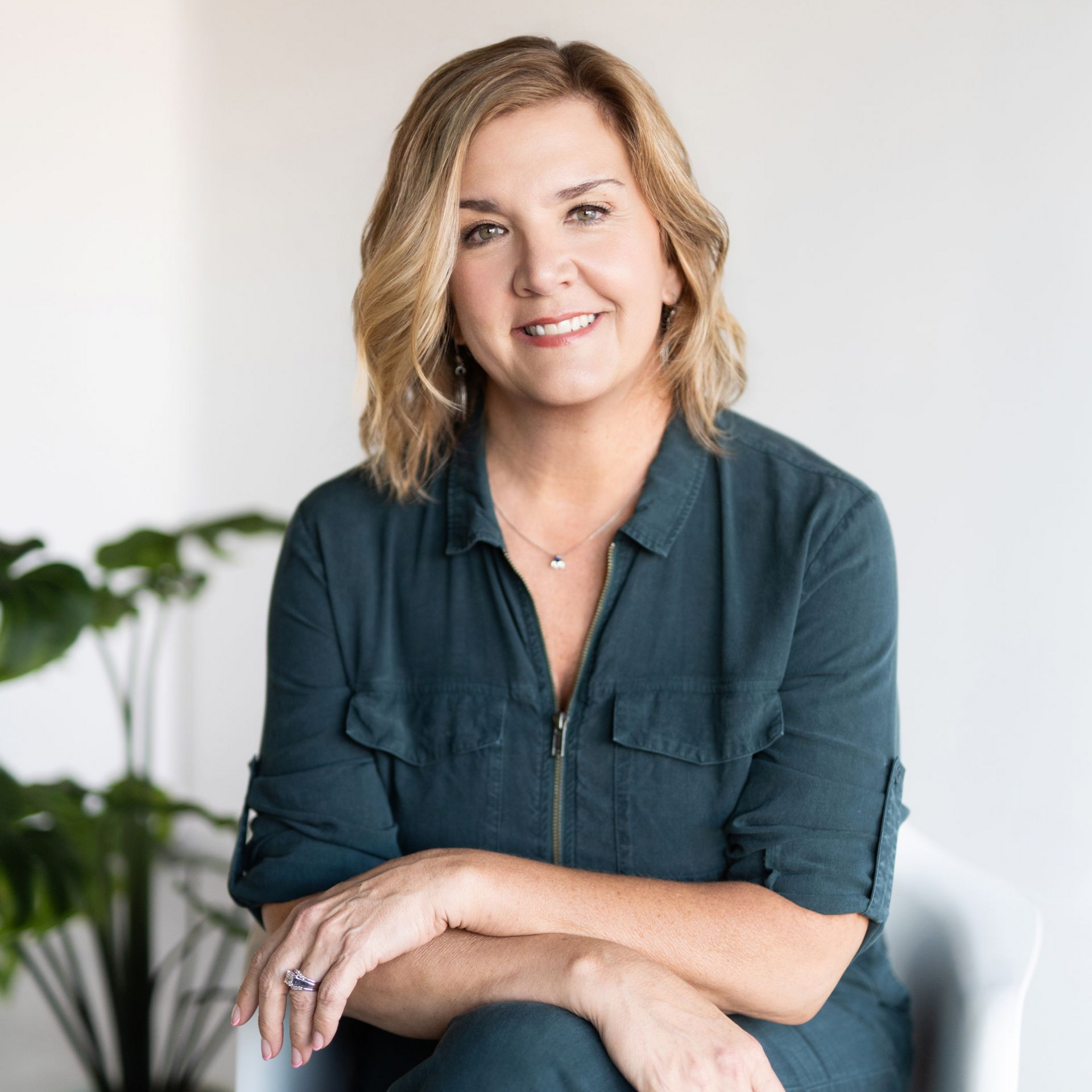 Portrait of a smiling person in a teal shirt seated indoors with arms crossed, near a plant