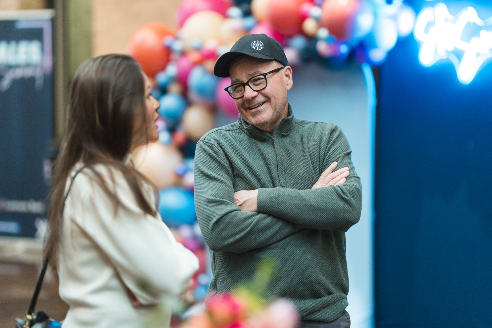 Man in cap and glasses smiles, arms crossed, talking to a woman. Balloons and neon sign in the background.