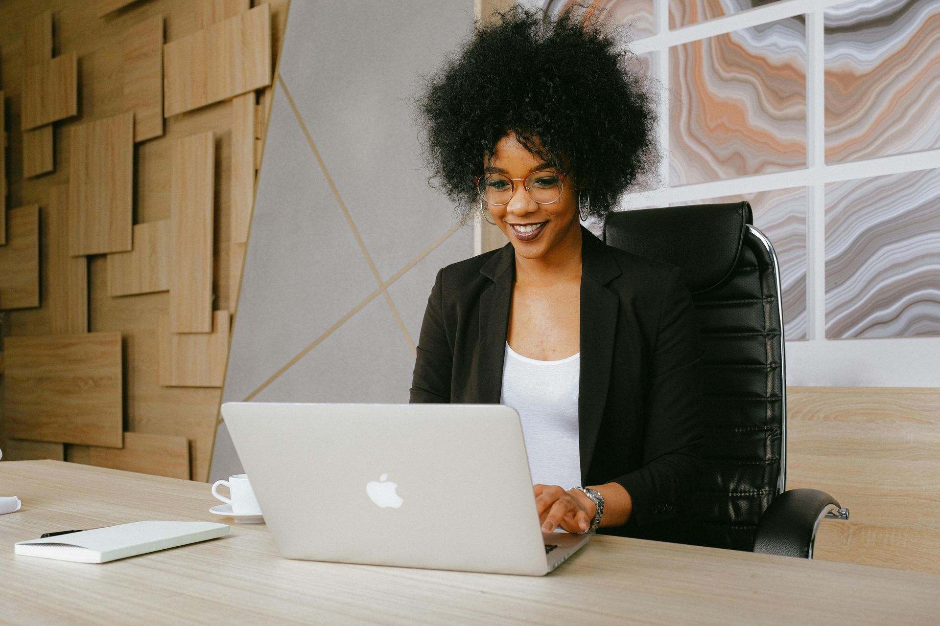 Woman with afro smiles while working on a laptop at a desk in an office.