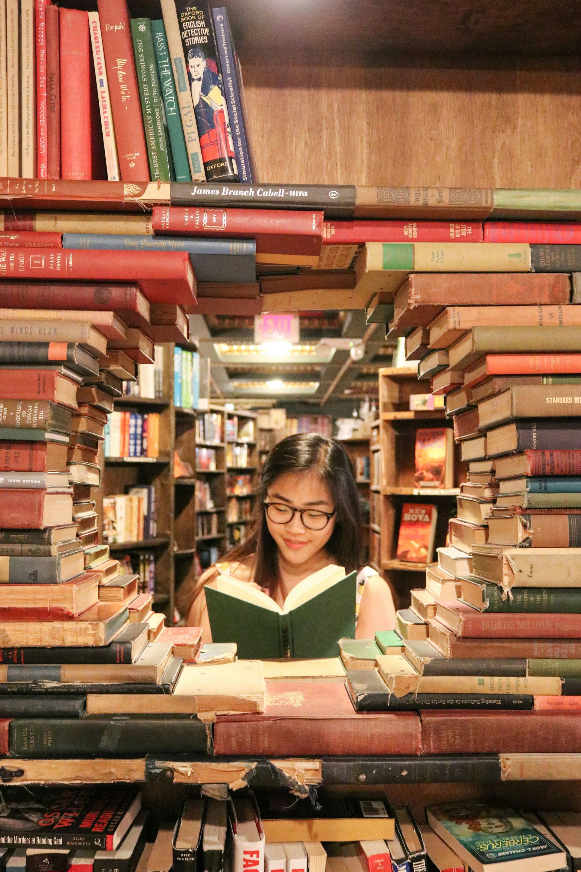 Uma jovem de óculos lê um livro cercada por pilhas de livros antigos em uma livraria.
