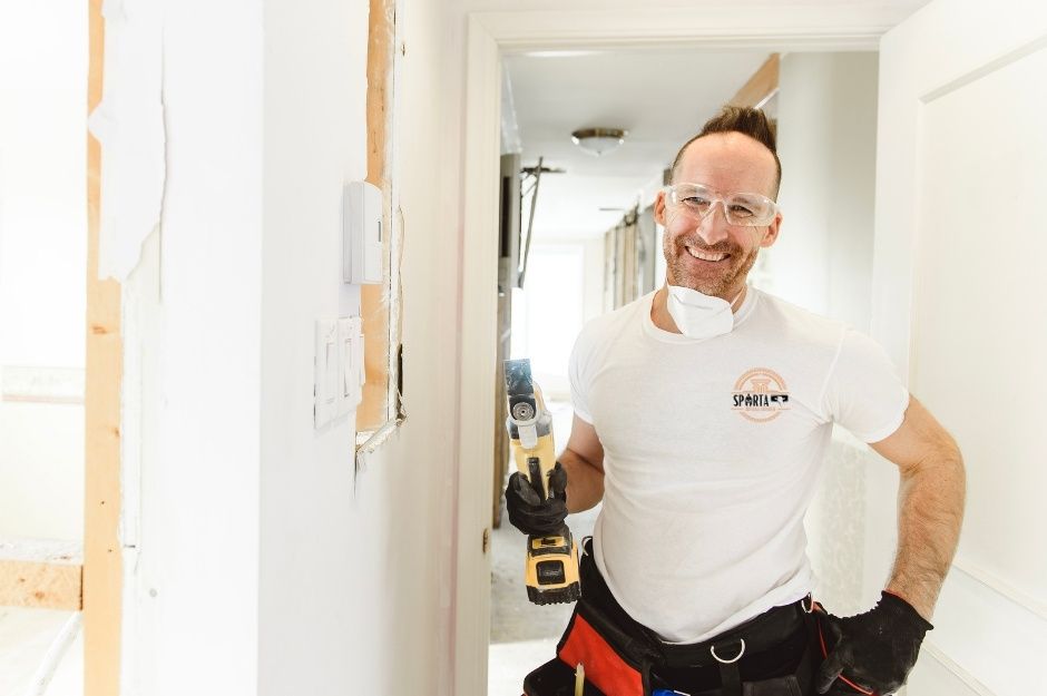A smiling drywall contractor wearing a white shirt stands with a power drill in hand next to completed drywall installation and wooden wall framing. This contractor is completing work near W Superstition Blvd, and N Pinyon Dr, Apache Junction, AZ.