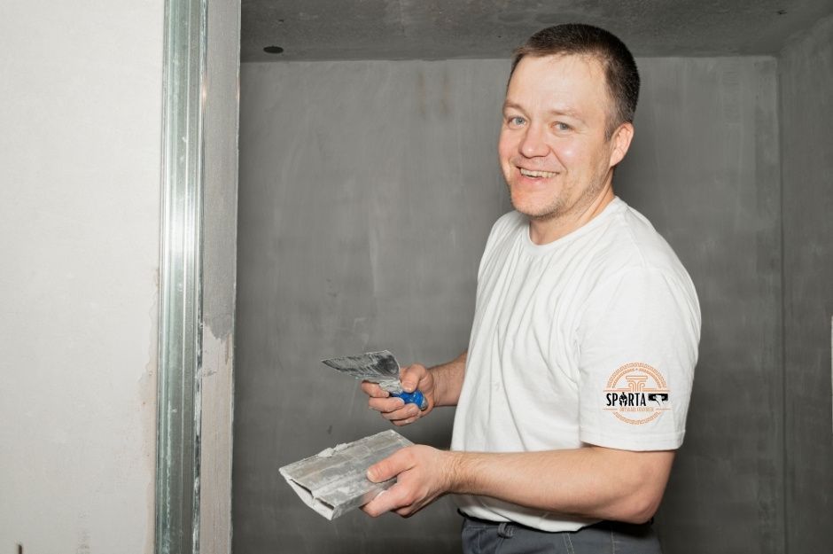 A drywall contractor in a white shirt holds joint compound application tools while working on interior wall finishing. The contractor is in the middle of a residential drywall installation project, with metal studs and drywall panels surrounding the work area near Lapidary Rd, and Chrysocolla Rd, Black Canyon, Arizona.