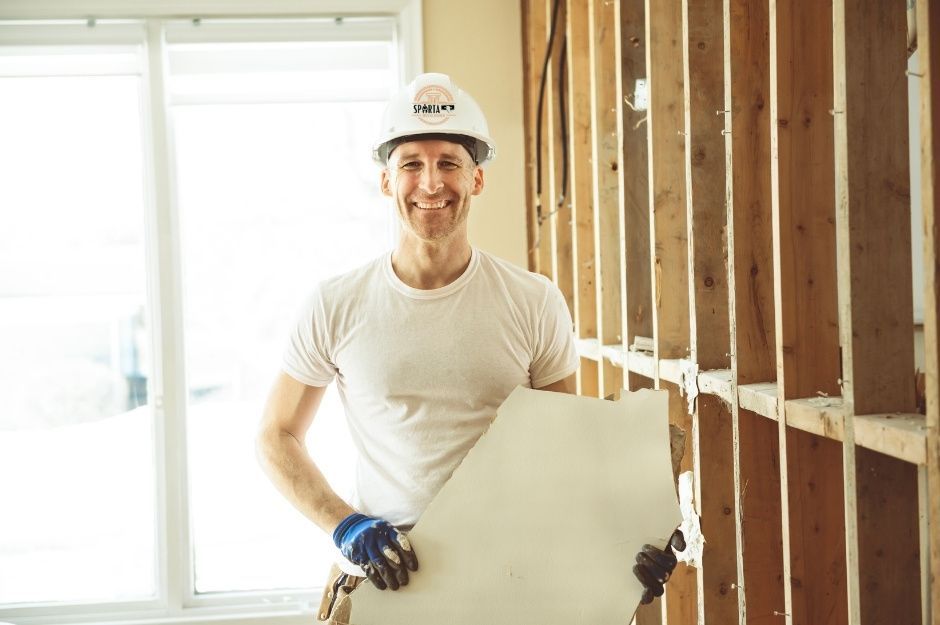 A drywall company worker in a white hard hat and light shirt holds a large white drywall sheet against wooden wall framing. He wears blue work gloves and stands in a bright residential construction site with exposed studs and natural lighting near W Vah Ki Inn Rd, and N 3rd St, Coolidge, AZ.