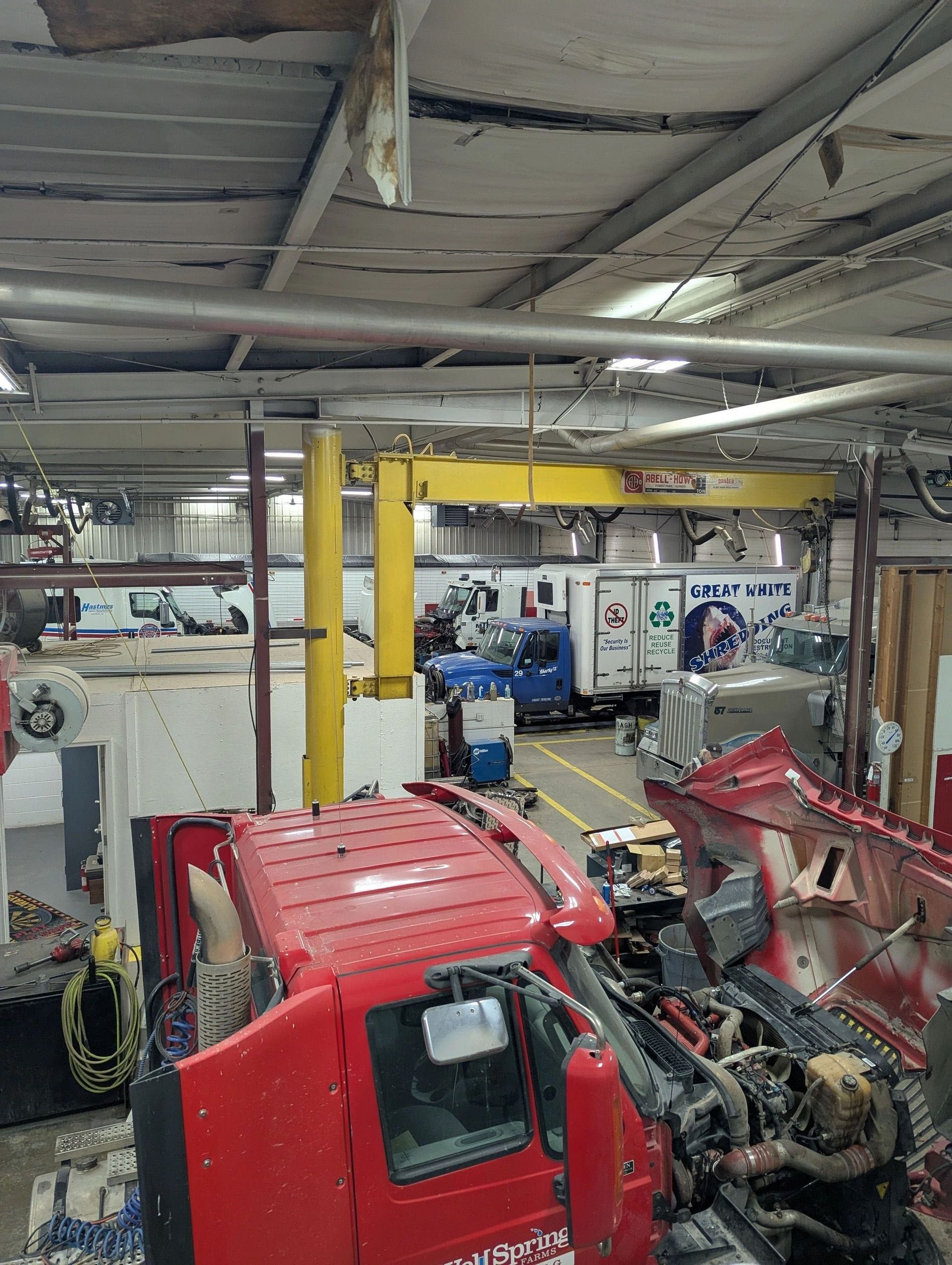 Red truck being worked on in a large garage. A yellow crane and several other trucks are visible in the background.