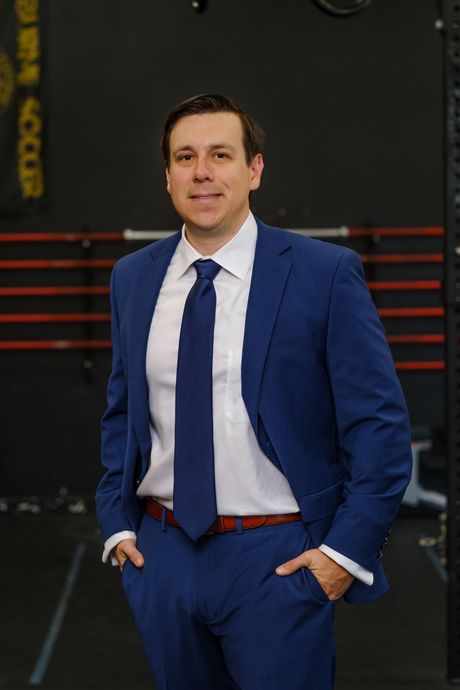A man in a blue suit and tie is standing in a gym with his hands in his pockets.
