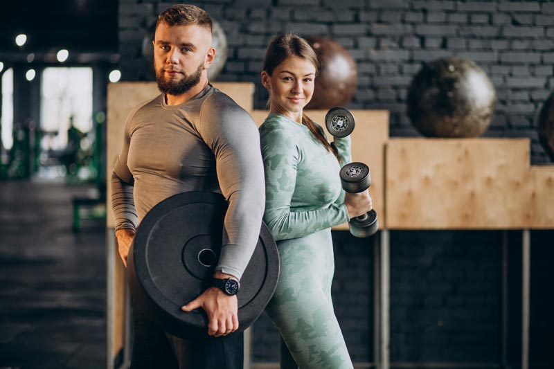 A man and a woman are standing back to back in a gym holding weights.