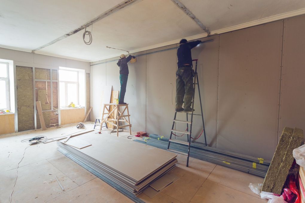 Renovation room with workers on ladders patching walls, drywall stacked on the floor.