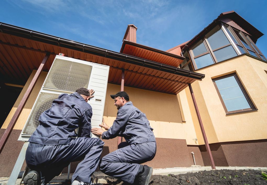 Two workers kneel beside a house exterior, examining a wall under the eaves.