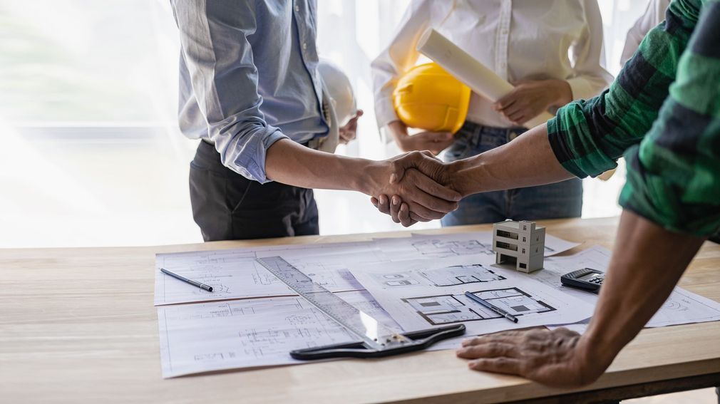 Colleagues in hard hats reviewing blueprint plans and shaking hands at a construction site table