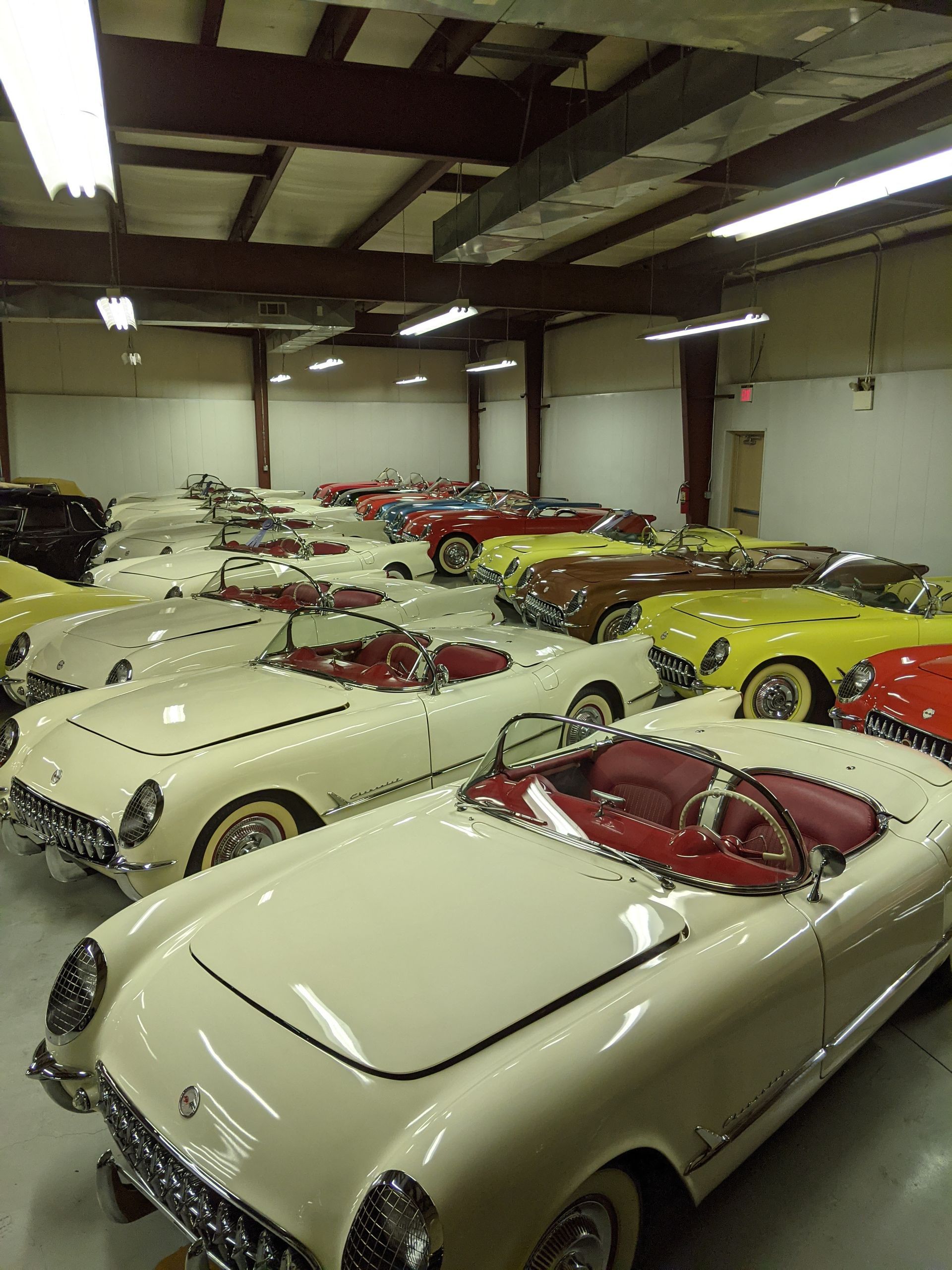 Rows of vintage sports cars in a garage, mostly cream-colored with red interiors and one bright yellow car.
