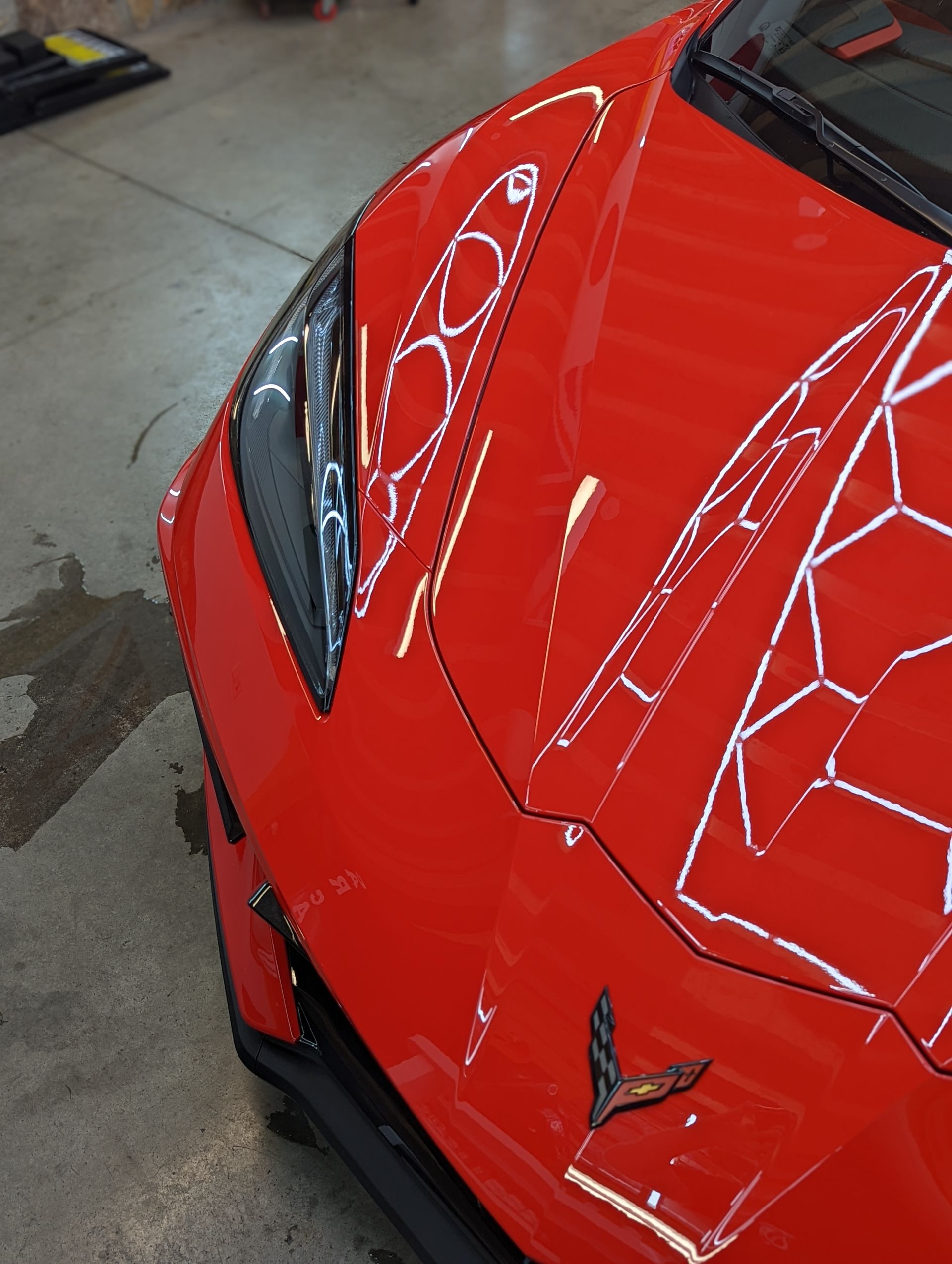 Close-up of a red sports car hood and headlight in a garage, with white reflection lines on the paint