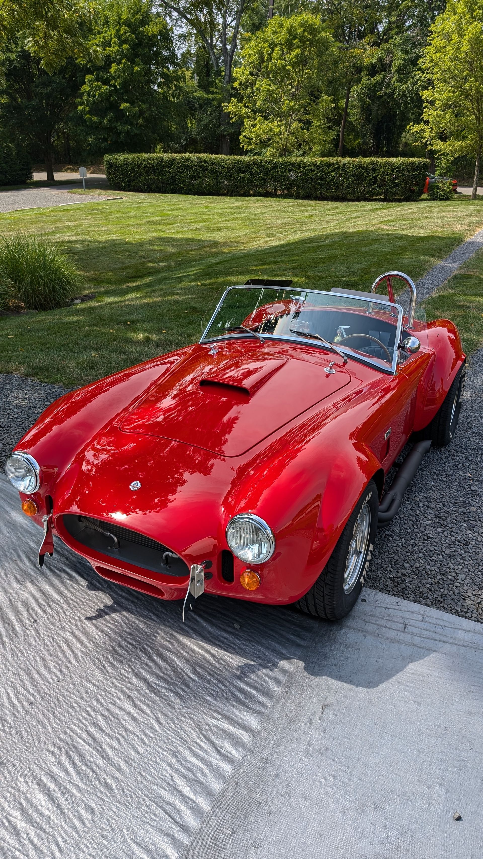 Red vintage convertible sports car parked on a gravel driveway beside a manicured lawn and trees