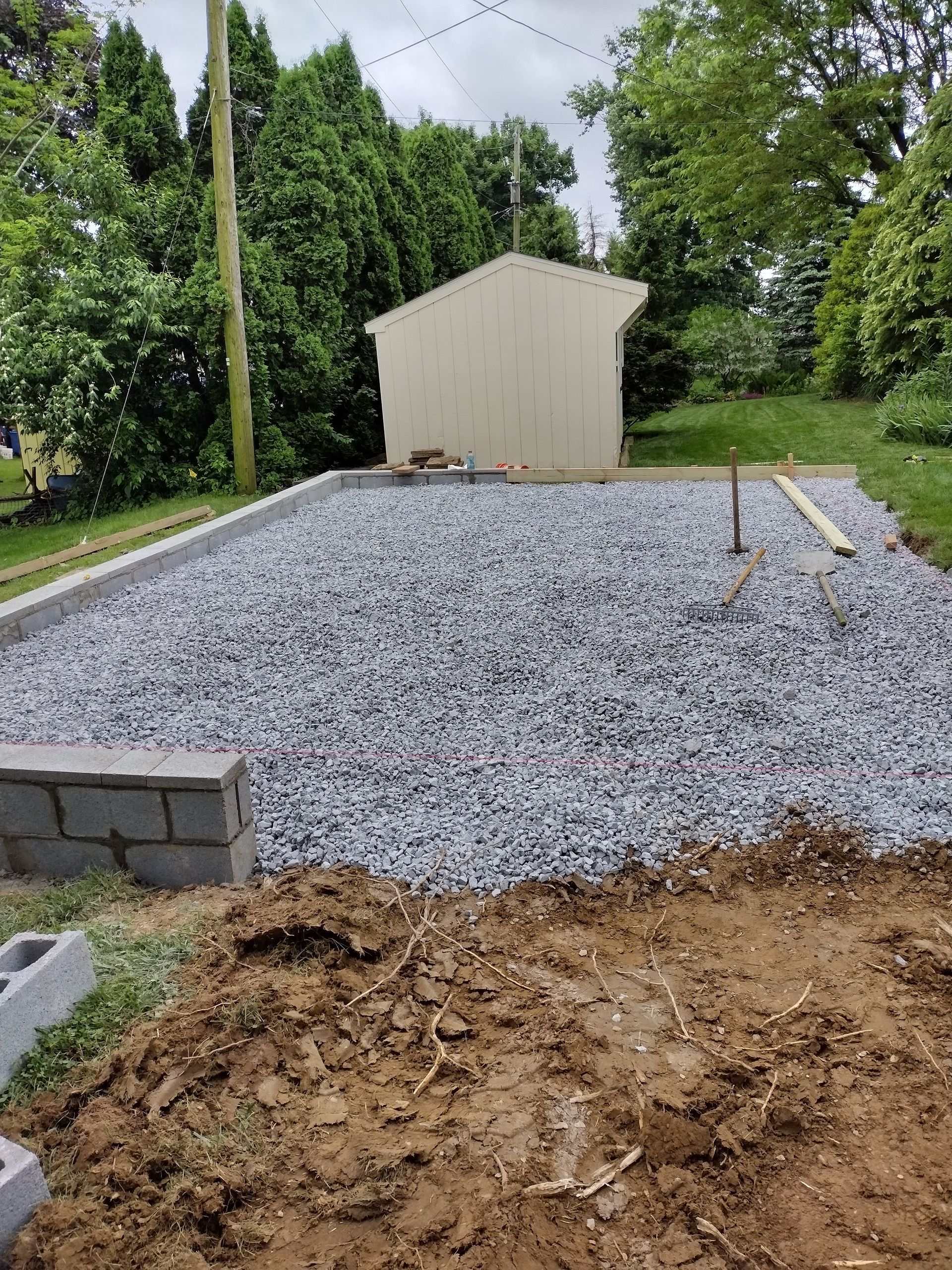 A backyard construction site featuring a gravel-filled foundation, concrete blocks, and a small shed in the background.