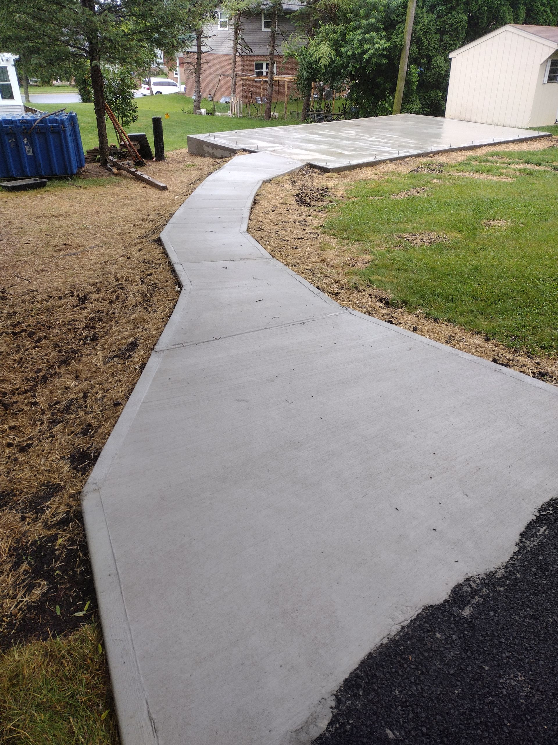 Newly poured concrete path leading from a black gravel area to a concrete patio in a backyard setting.