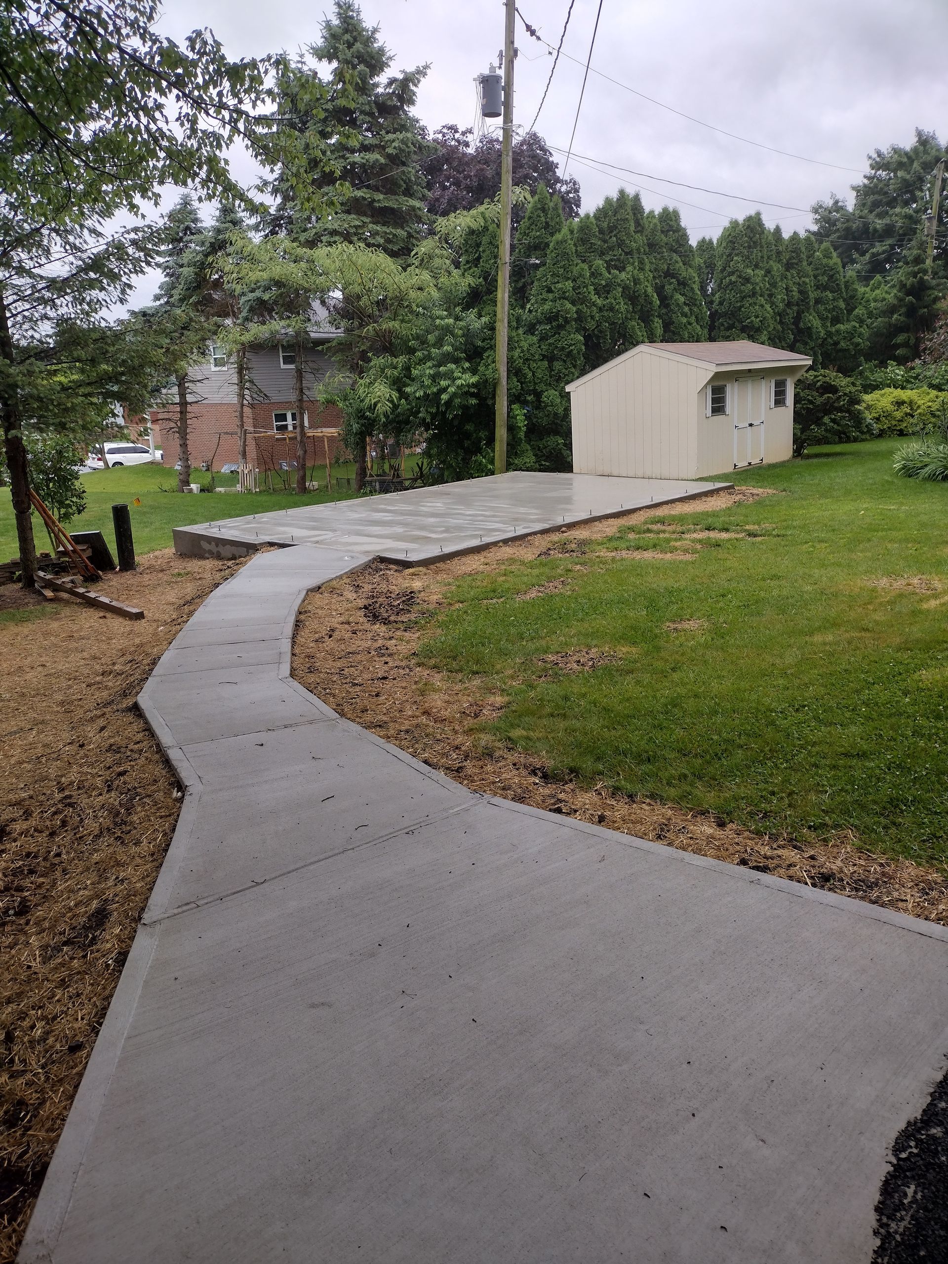 A concrete walkway leads through a grassy yard to a small, light-colored storage shed under a cloudy sky.