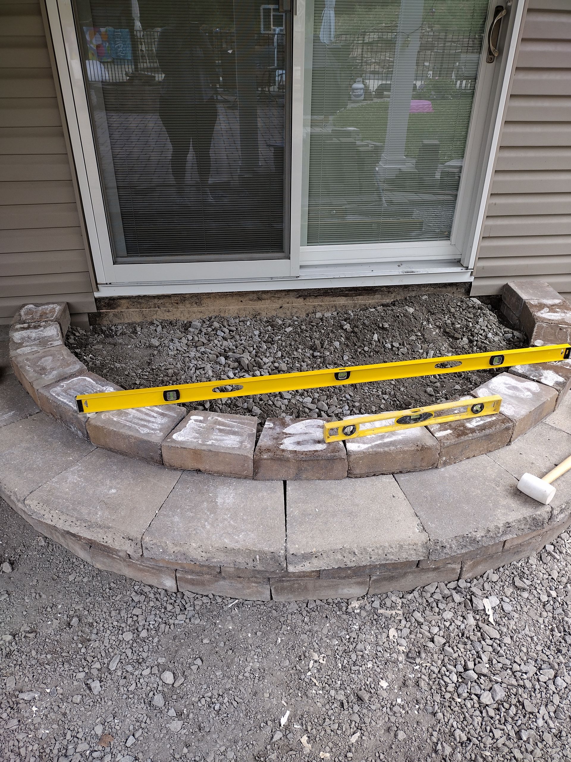 A partially built curved stone step outside a sliding glass door, with two yellow leveling tools placed on top.