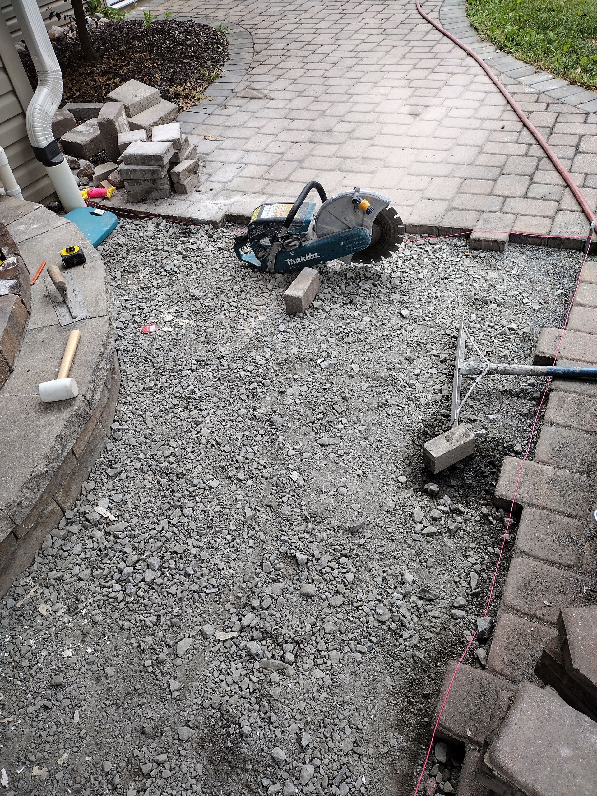 A power saw and tools sit on a gravel-filled walkway under construction, surrounded by unfinished brick pavers.
