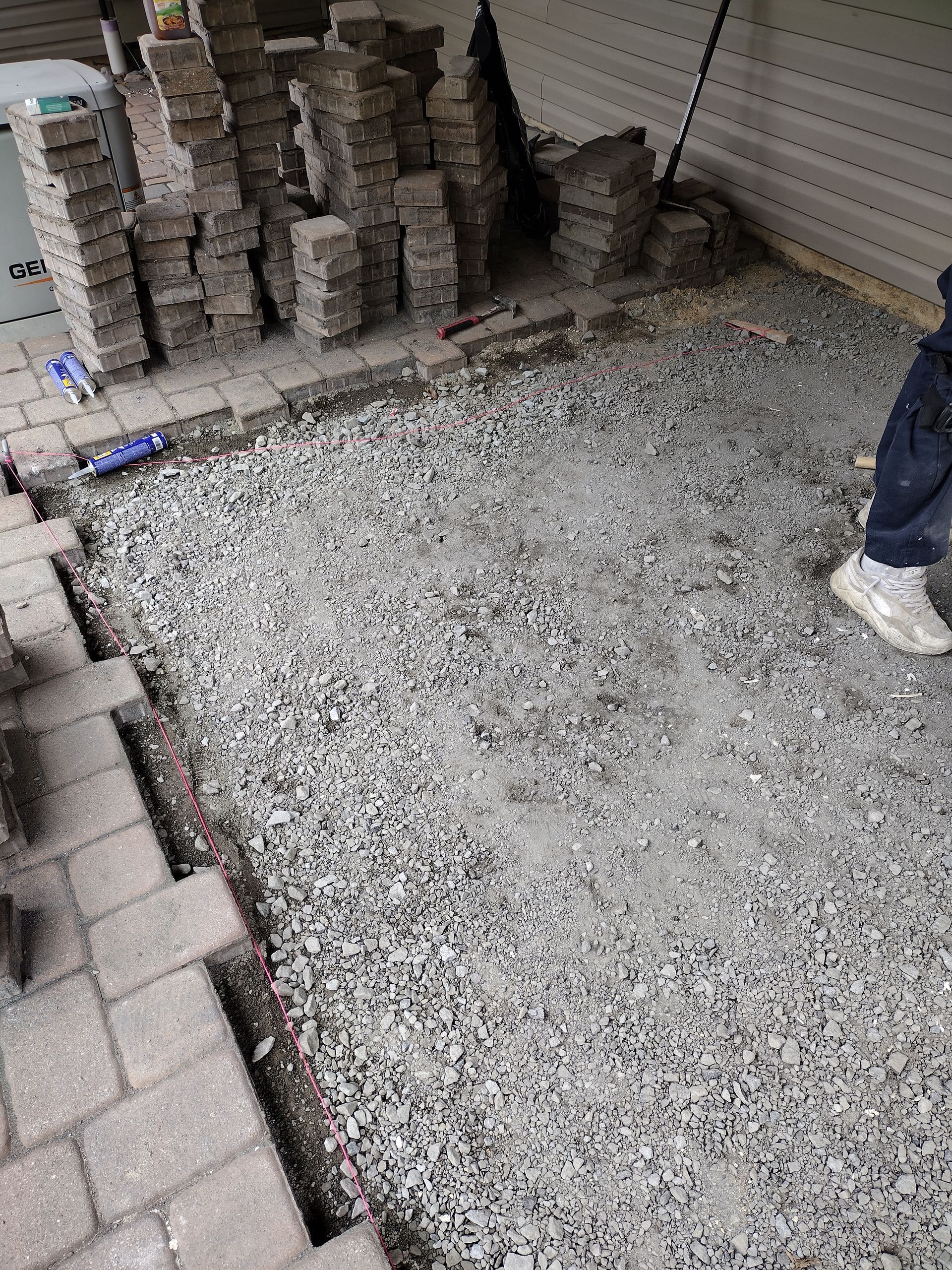 A gravel-filled rectangular area framed by laid stone pavers, with stacks of additional pavers nearby on a patio.