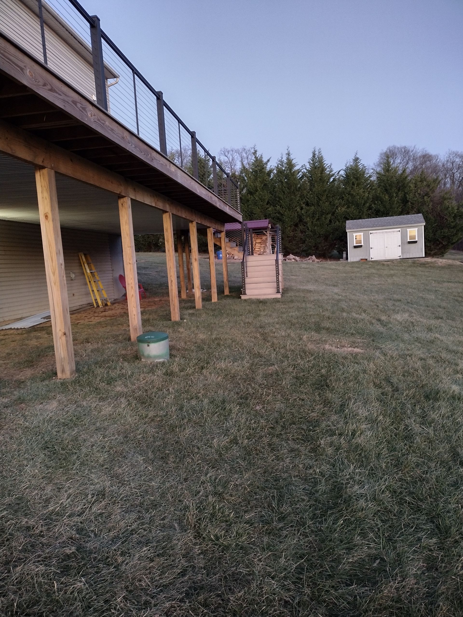 A low-angle view of a wooden deck with railing, stairs leading to the yard, and a shed in the background at dusk.