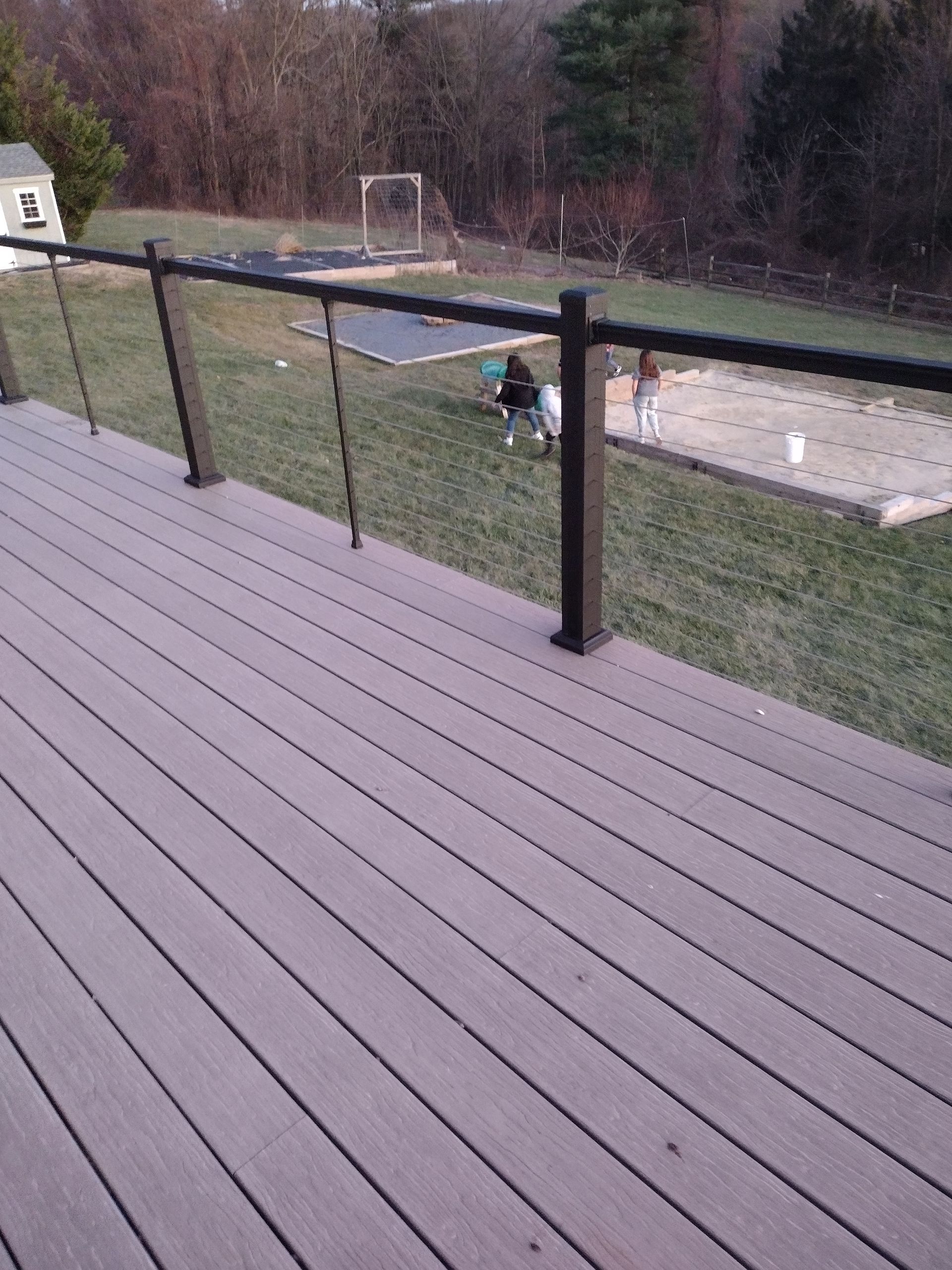 A view from a wooden deck looking out over a backyard where two people are standing in a sandy area near a garden.