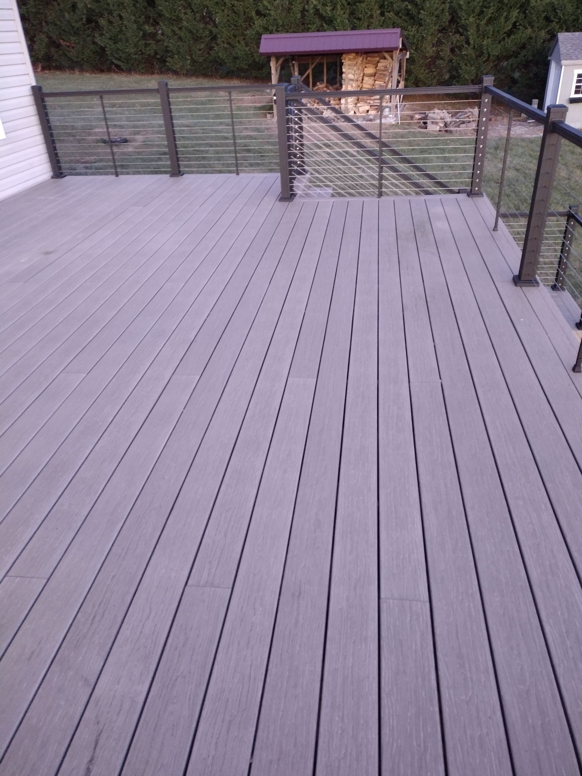 Gray composite deck with a metal cable railing, overlooking a grassy backyard with a small wooden structure.