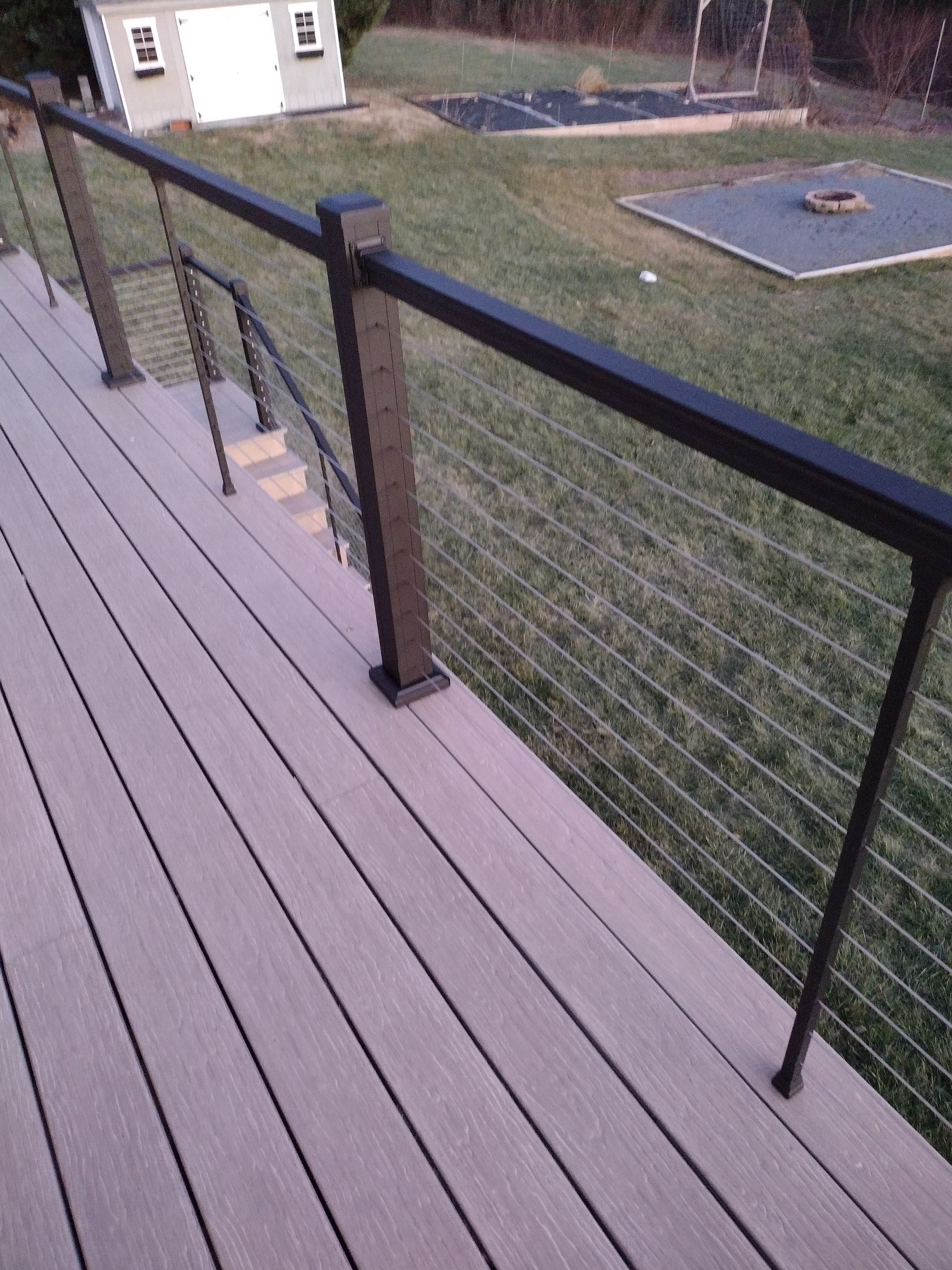 A view from a wooden deck looking over a backyard with a dark metal railing and thin horizontal cable infill.