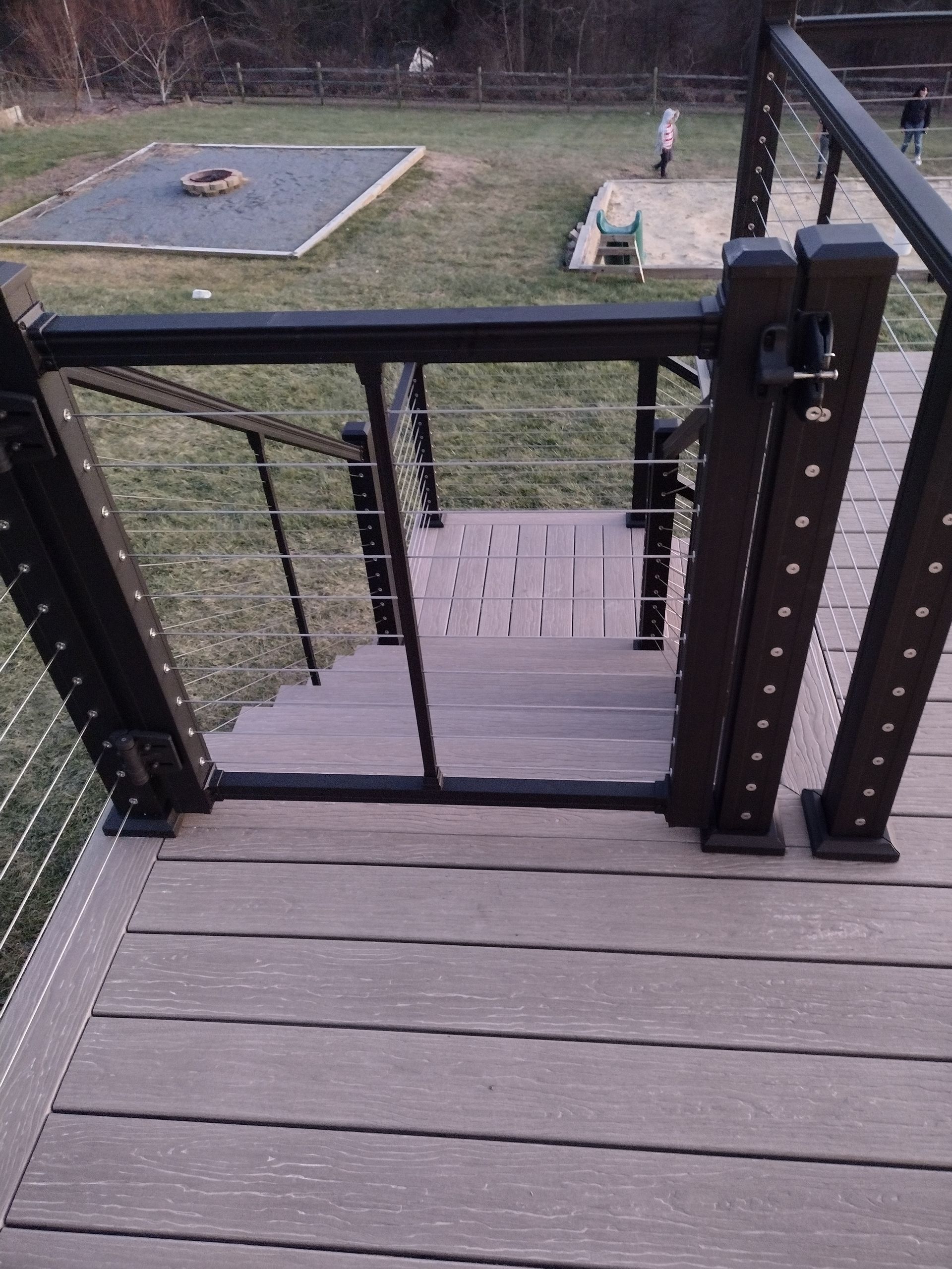 A black metal safety gate at the top of a composite deck staircase, overlooking a backyard with a fire pit.