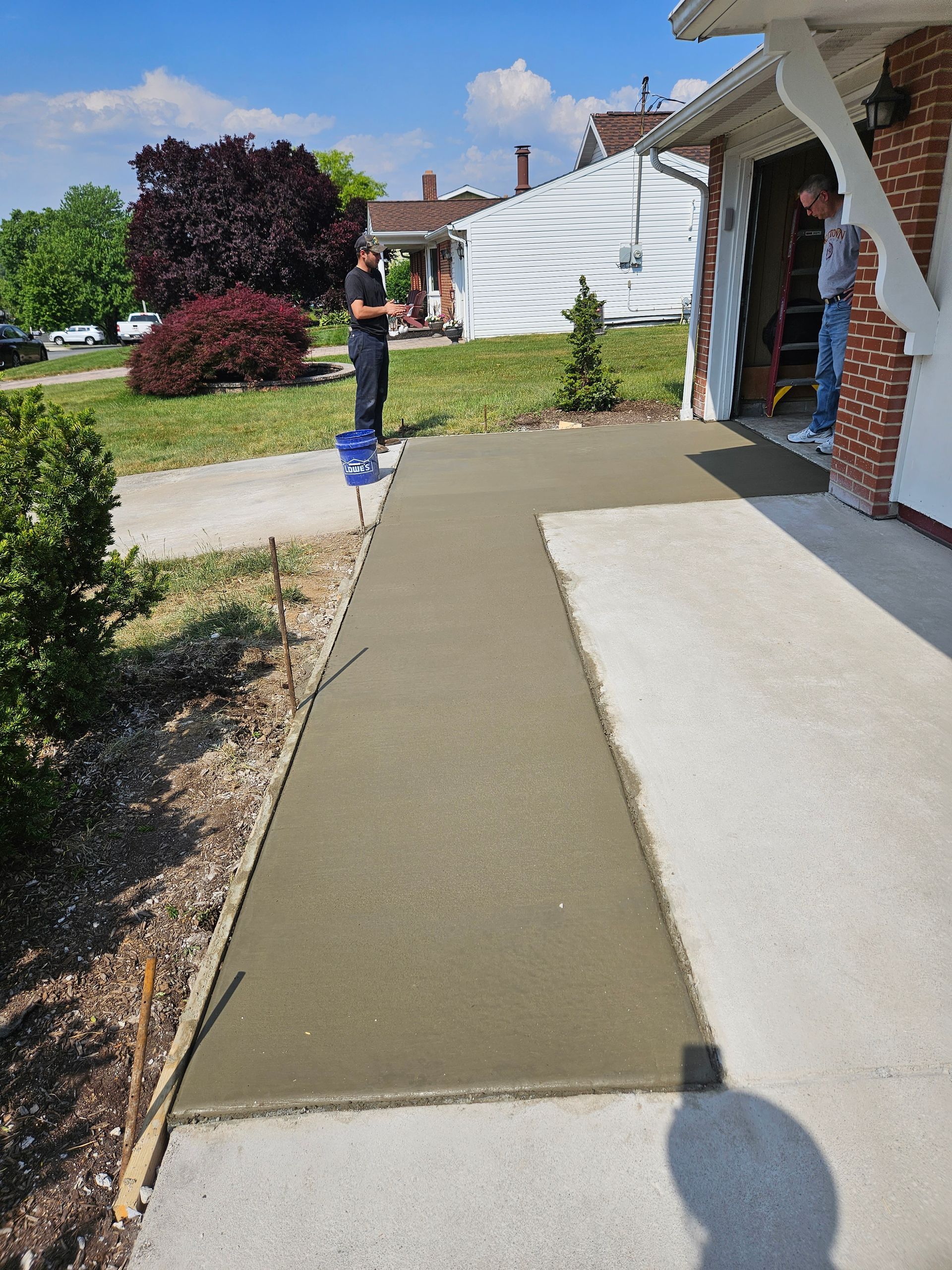 A newly poured concrete walkway extends from a house entrance toward a driveway, with two people standing nearby.