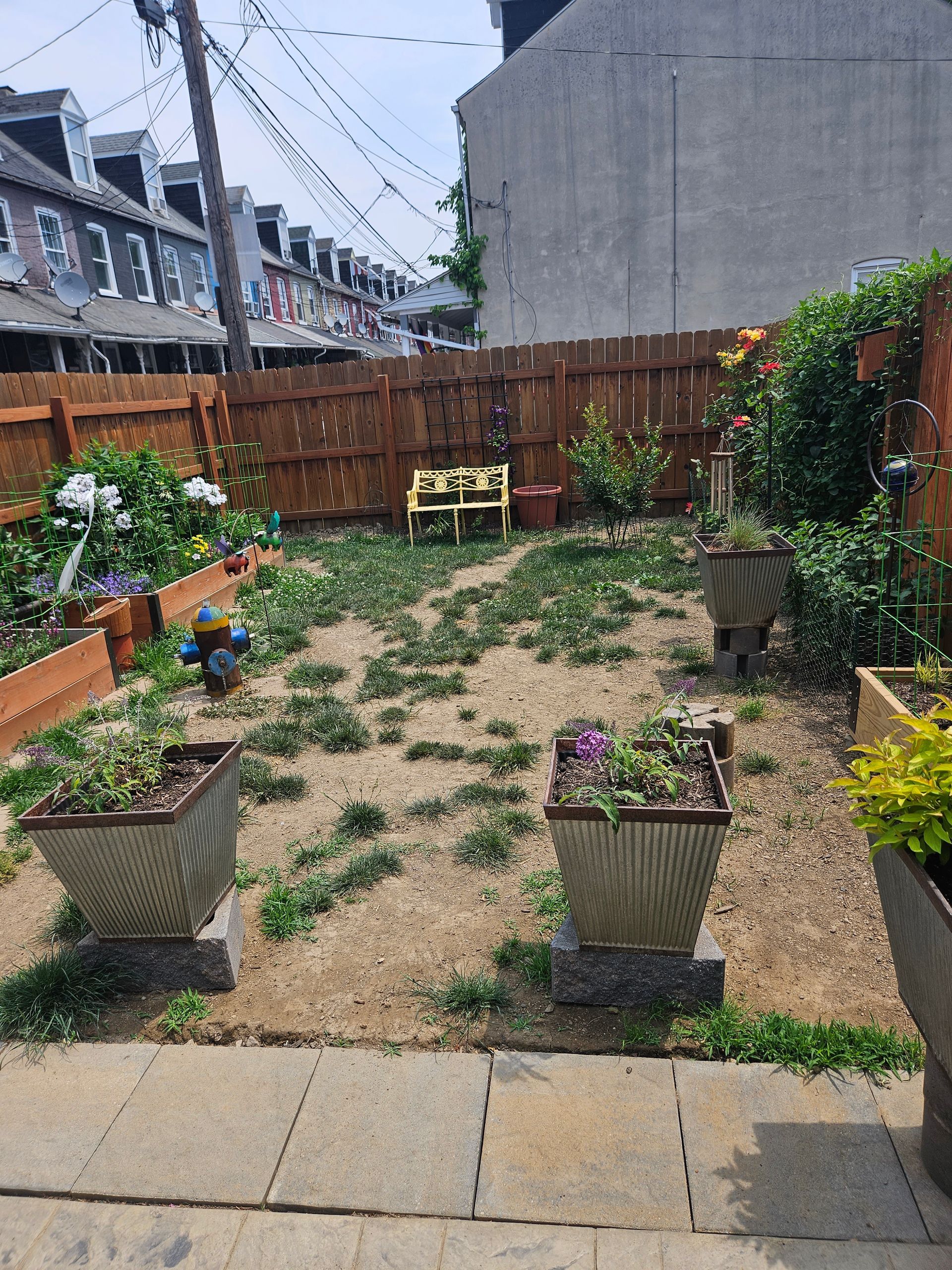 A small backyard garden featuring gravel ground with weeds, raised planter boxes, potted plants, and a wooden fence.