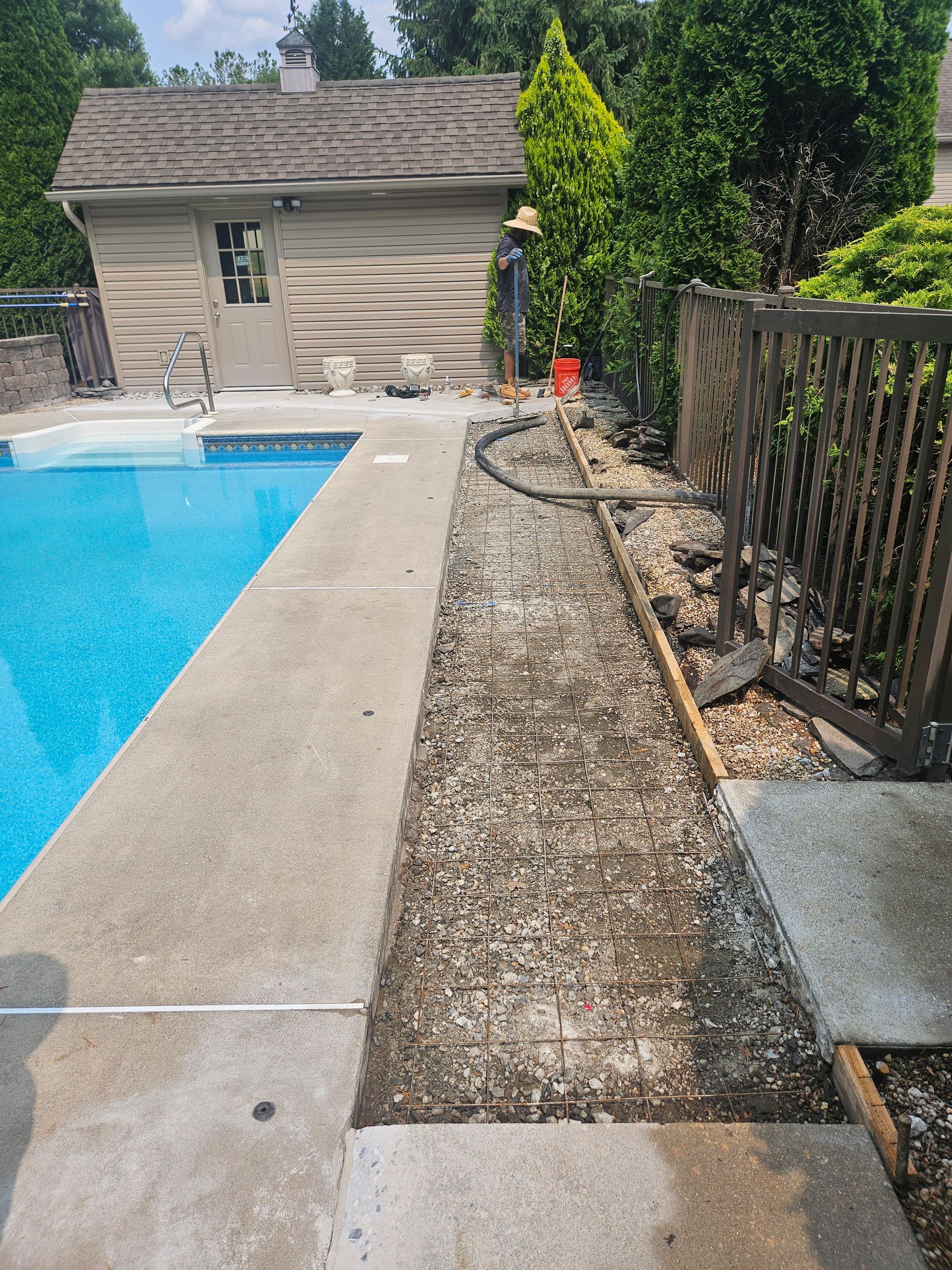 A backyard pool deck with a gravel-filled trench running alongside the concrete edge near a wooden fence and a small shed.