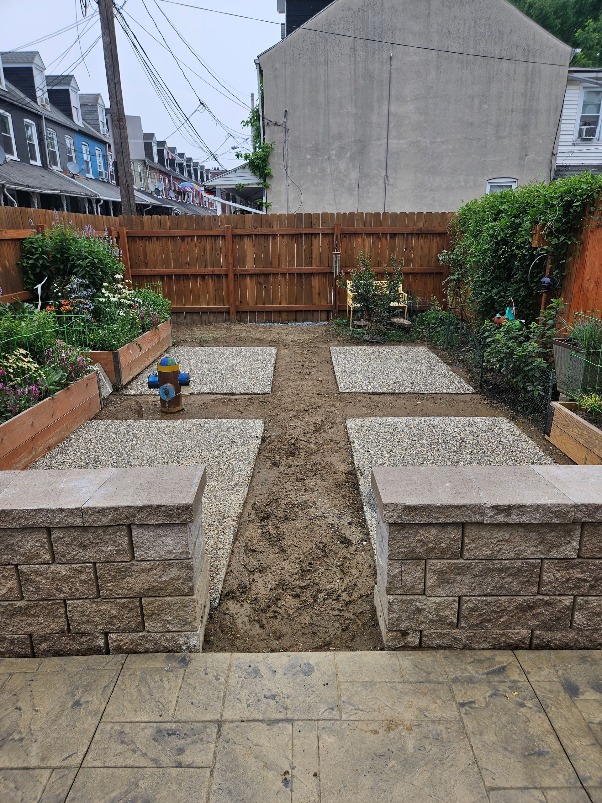 A backyard garden featuring four gravel squares, a central dirt path, raised wooden beds, and a stone retaining wall.
